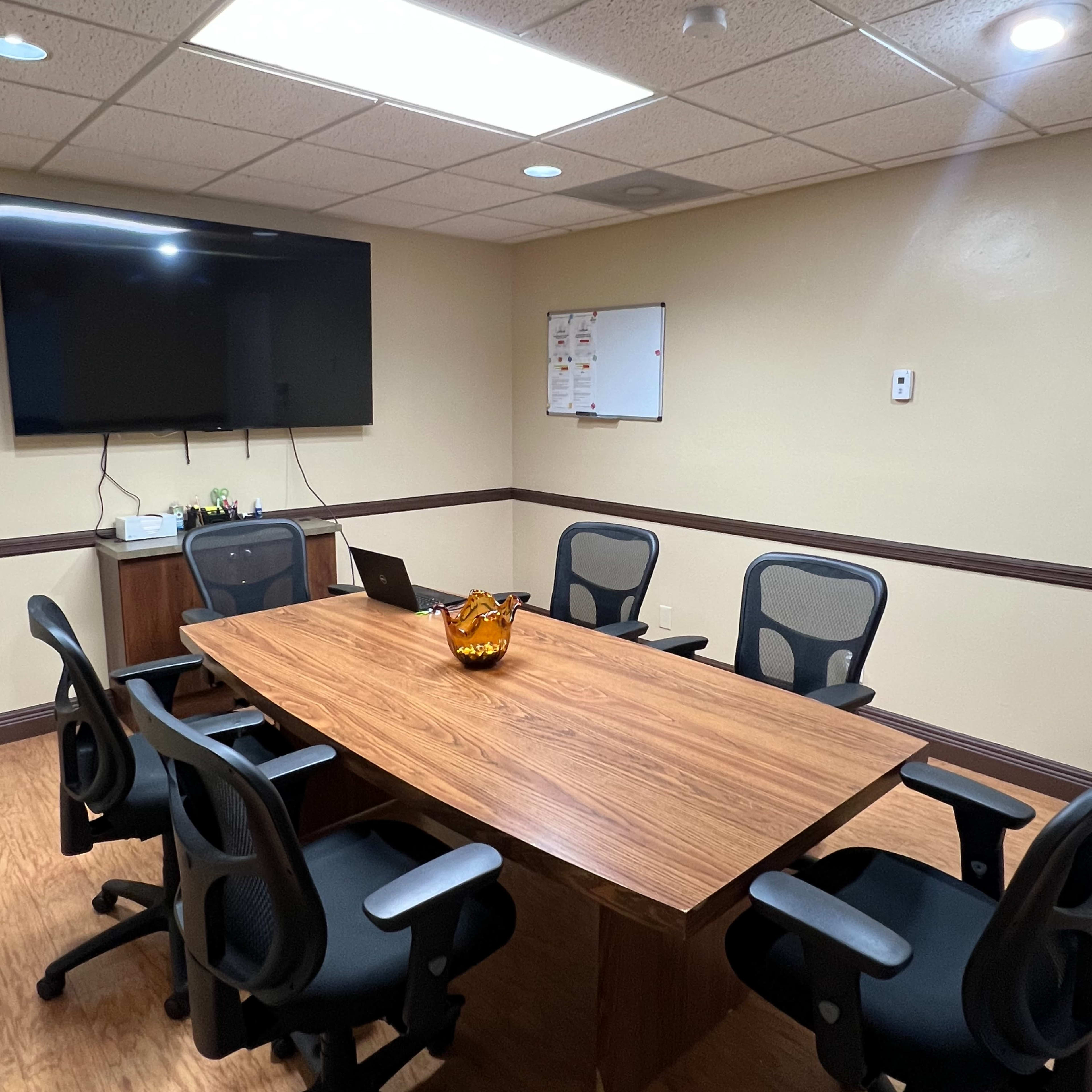 A conference room with a wooden table, six black office chairs, a television on the wall, and a whiteboard.