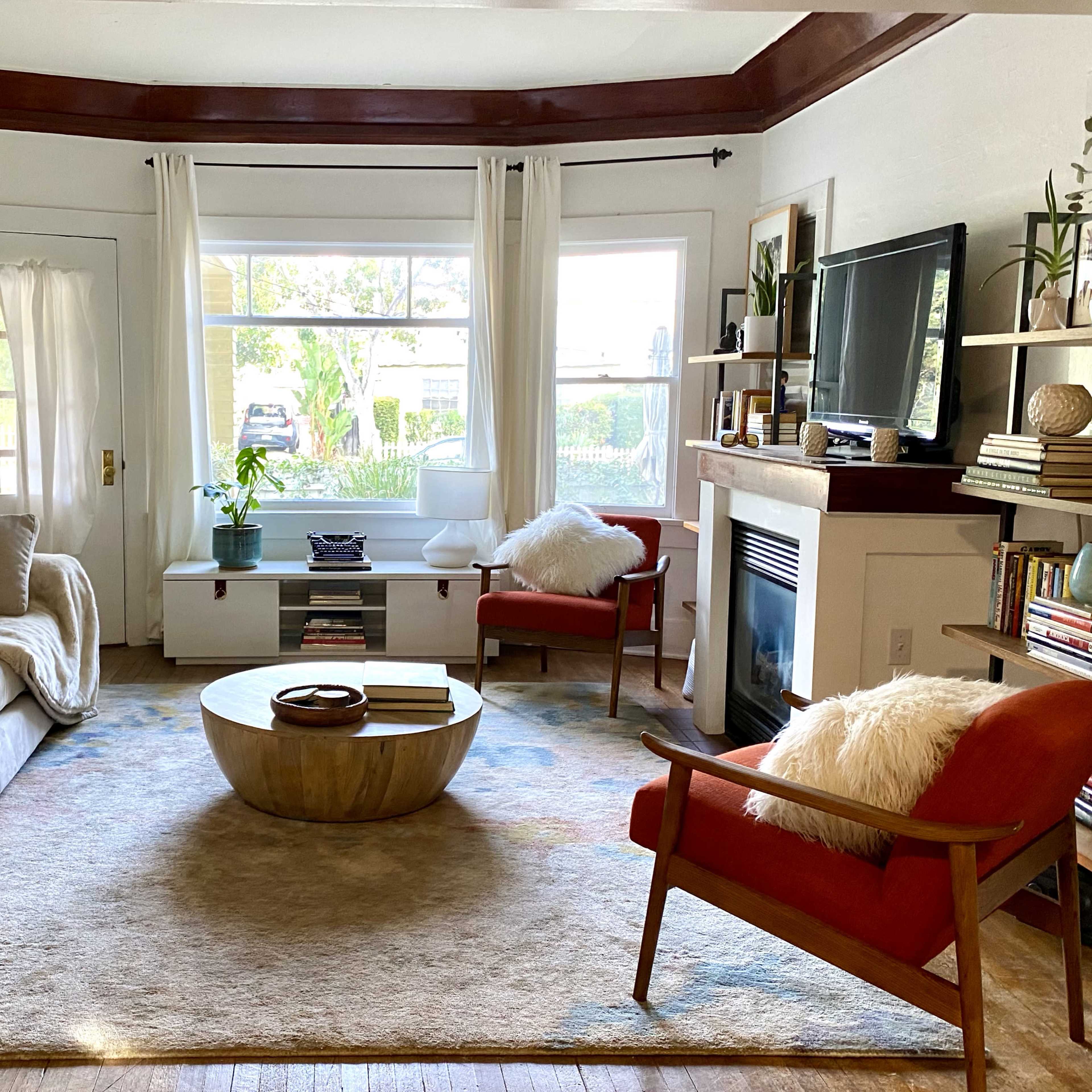 A cozy living room with a light gray sofa, two orange chairs, a round wooden coffee table, and large windows that provide natural light.
