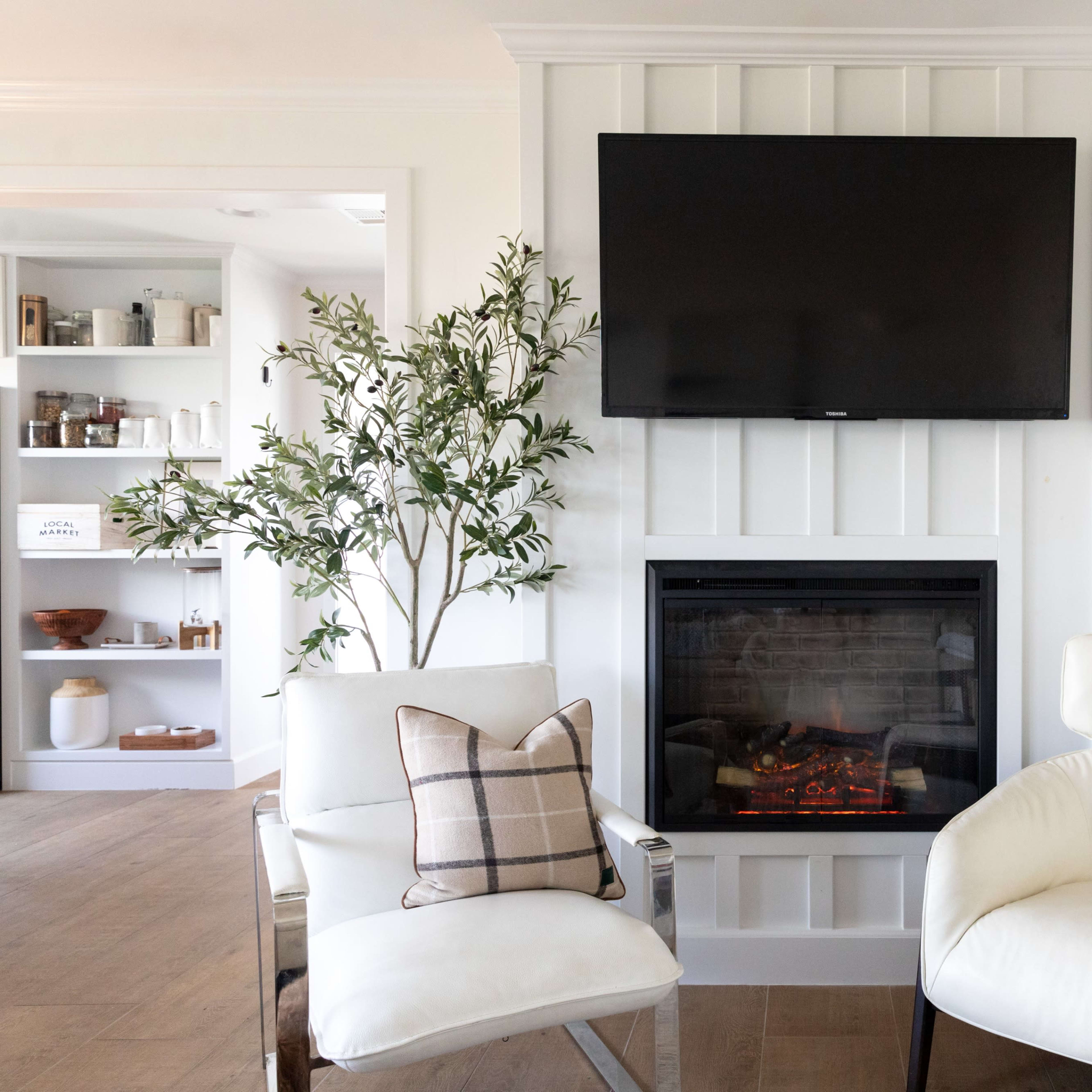 A living room with two white armchairs, a television mounted on a wall above a fireplace, and a decorative plant on the side.