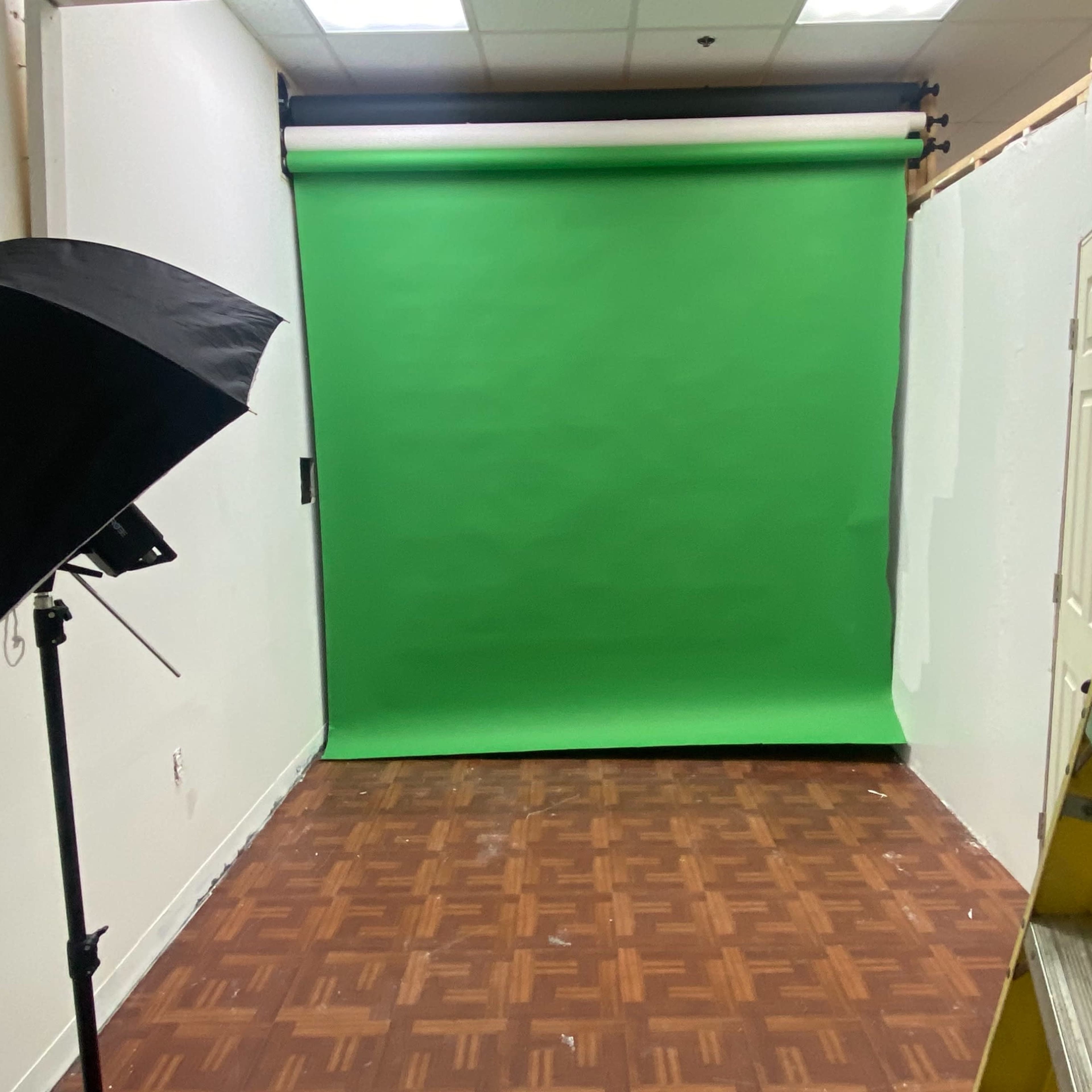 A small room with a green screen backdrop, a black umbrella light, and a ladder positioned against the wall.