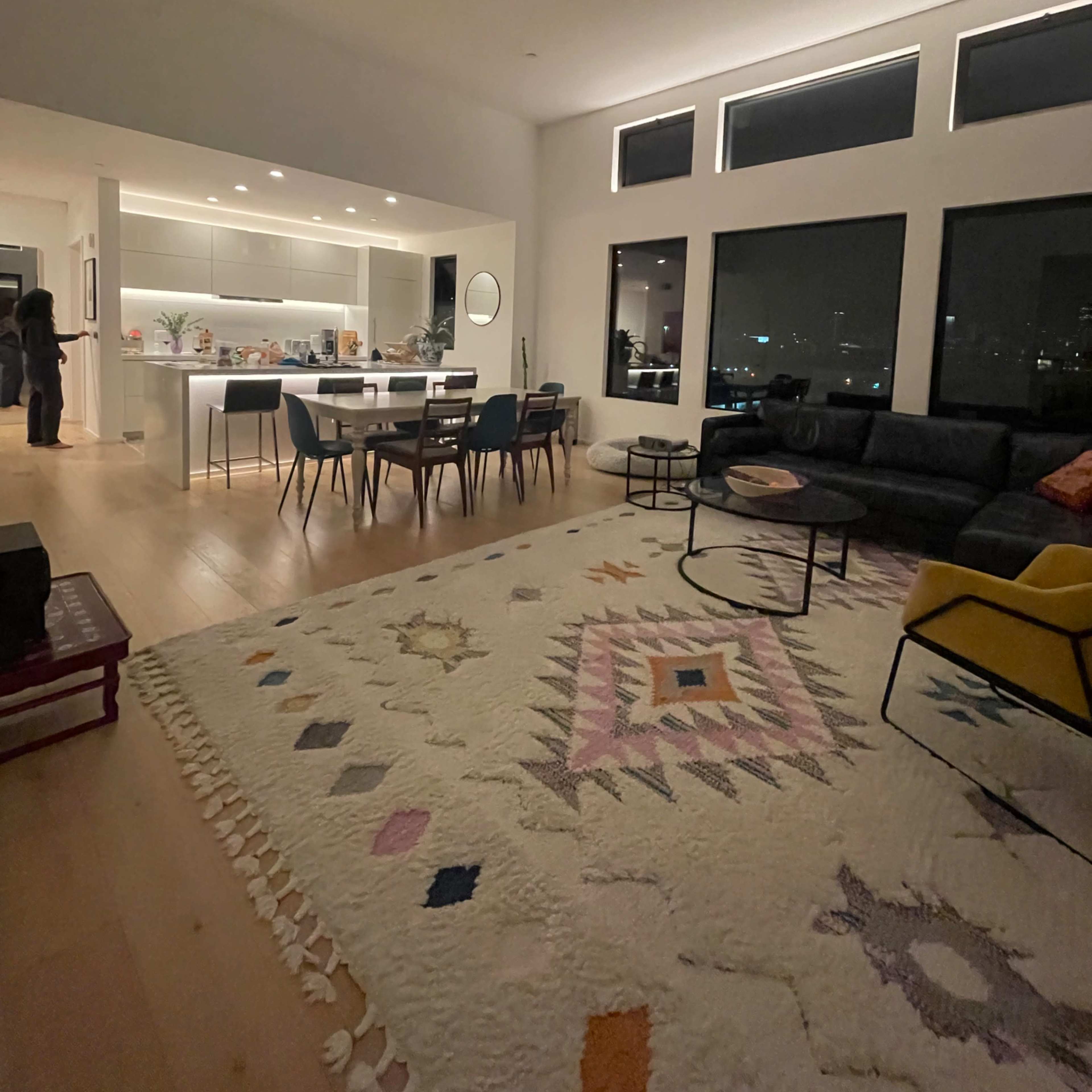 A modern living room features a large rug, a sectional sofa, a dining area with a table and chairs, and large windows overlooking a city view.