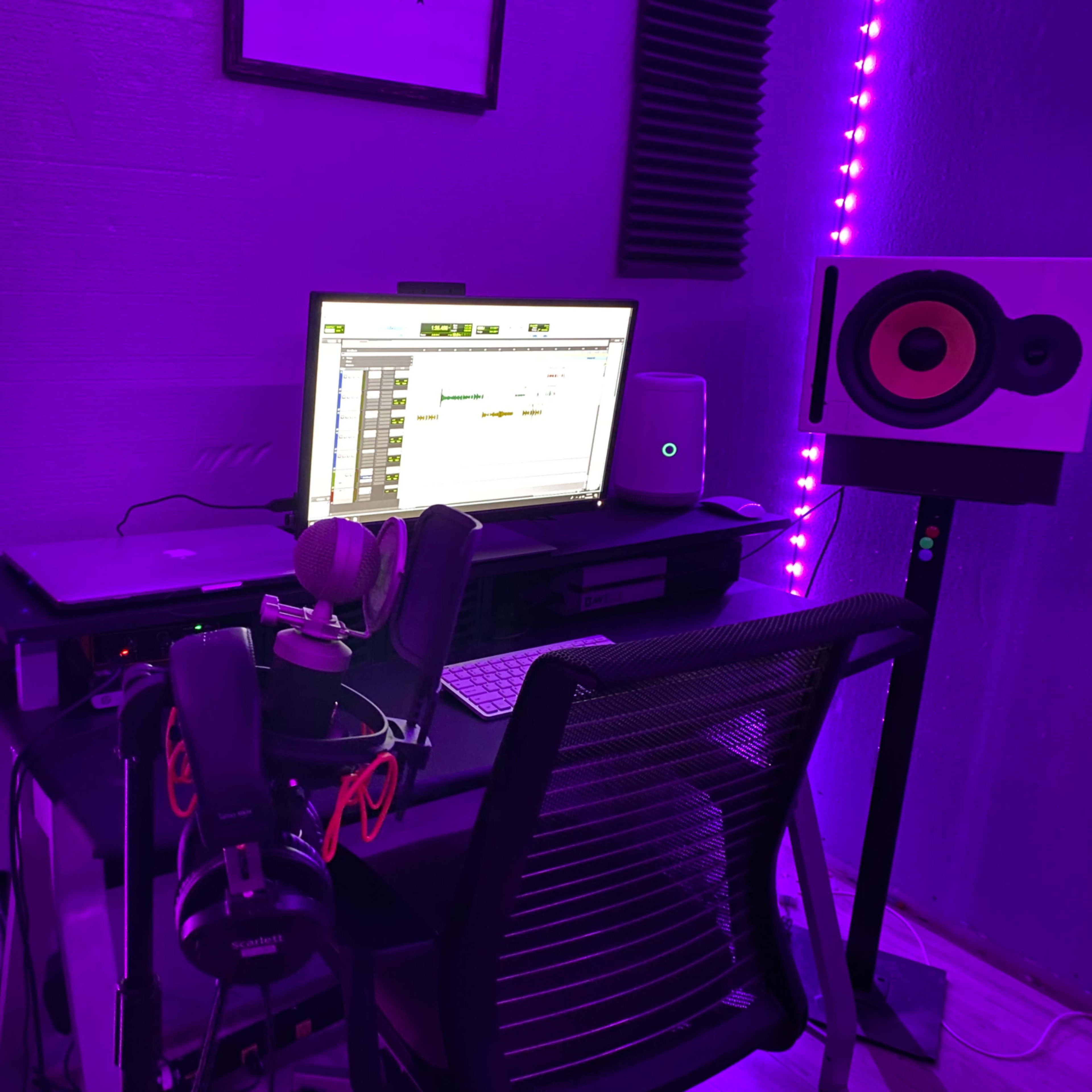 A home recording studio setup features a desk with a computer, audio interface, microphones, headphones, and two speakers, all illuminated by purple lighting.