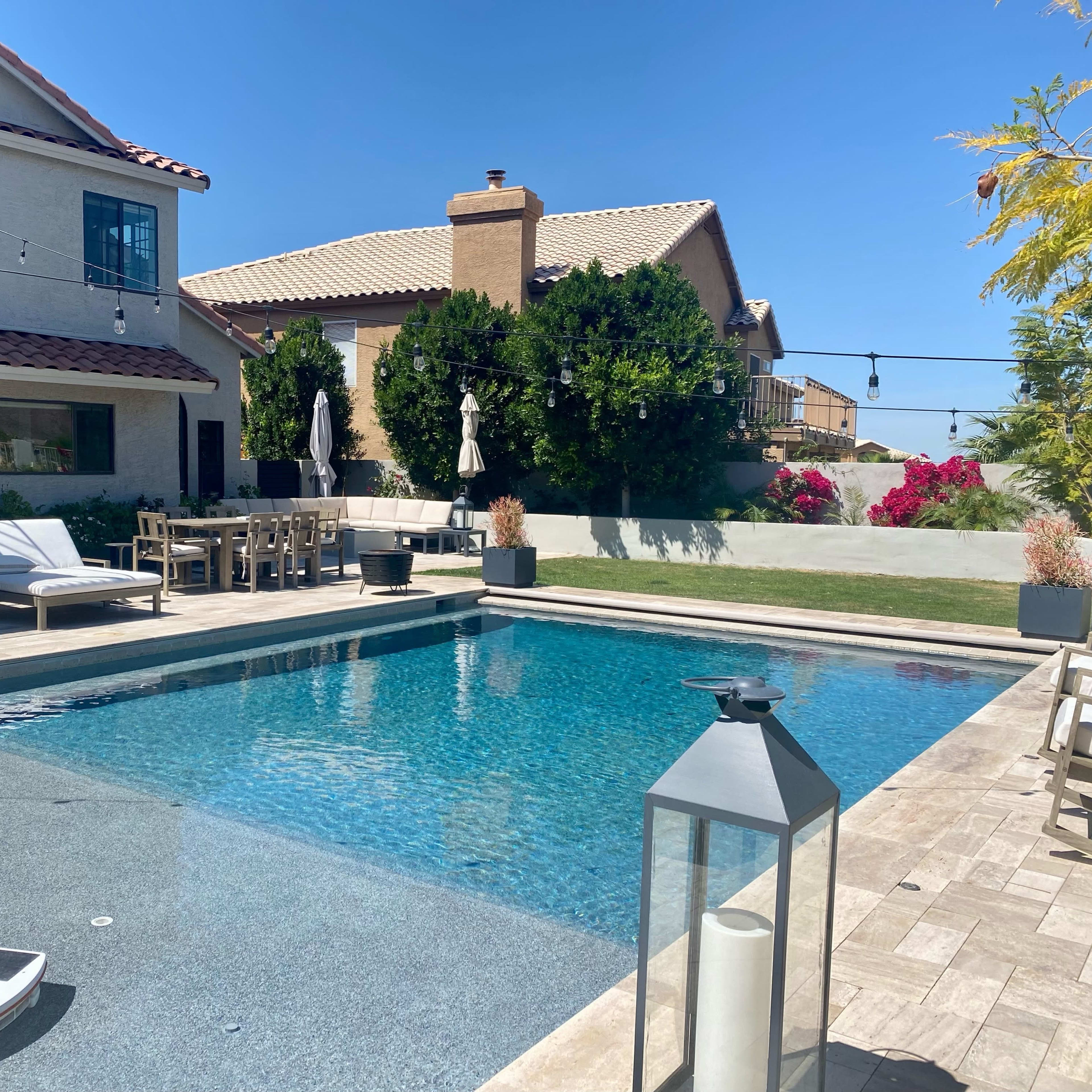 A backyard pool area featuring a clear swimming pool, lounge chairs, a dining table, and surrounding houses with landscaped vegetation.