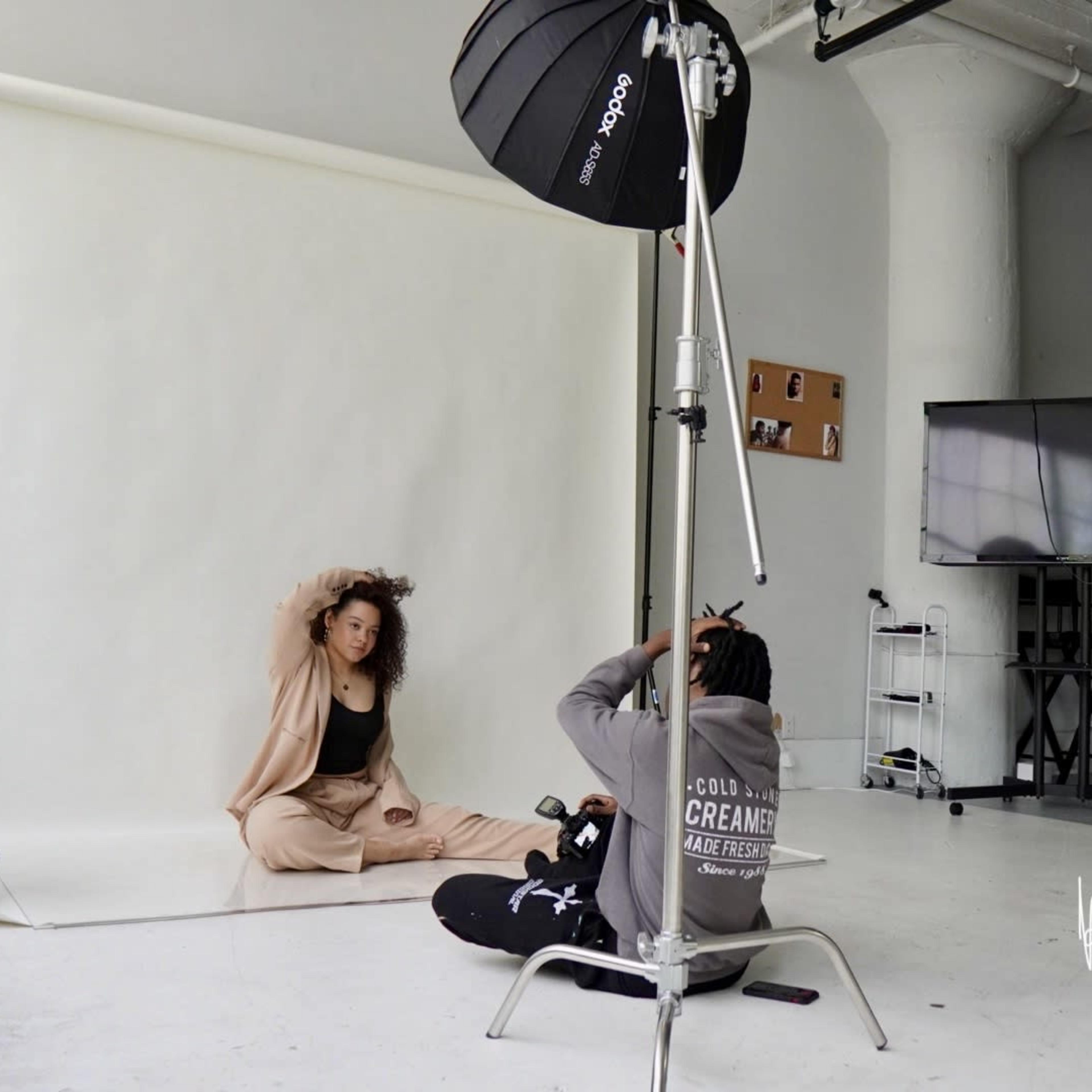 A model poses in front of a backdrop in a photography studio while a photographer adjusts the lighting.