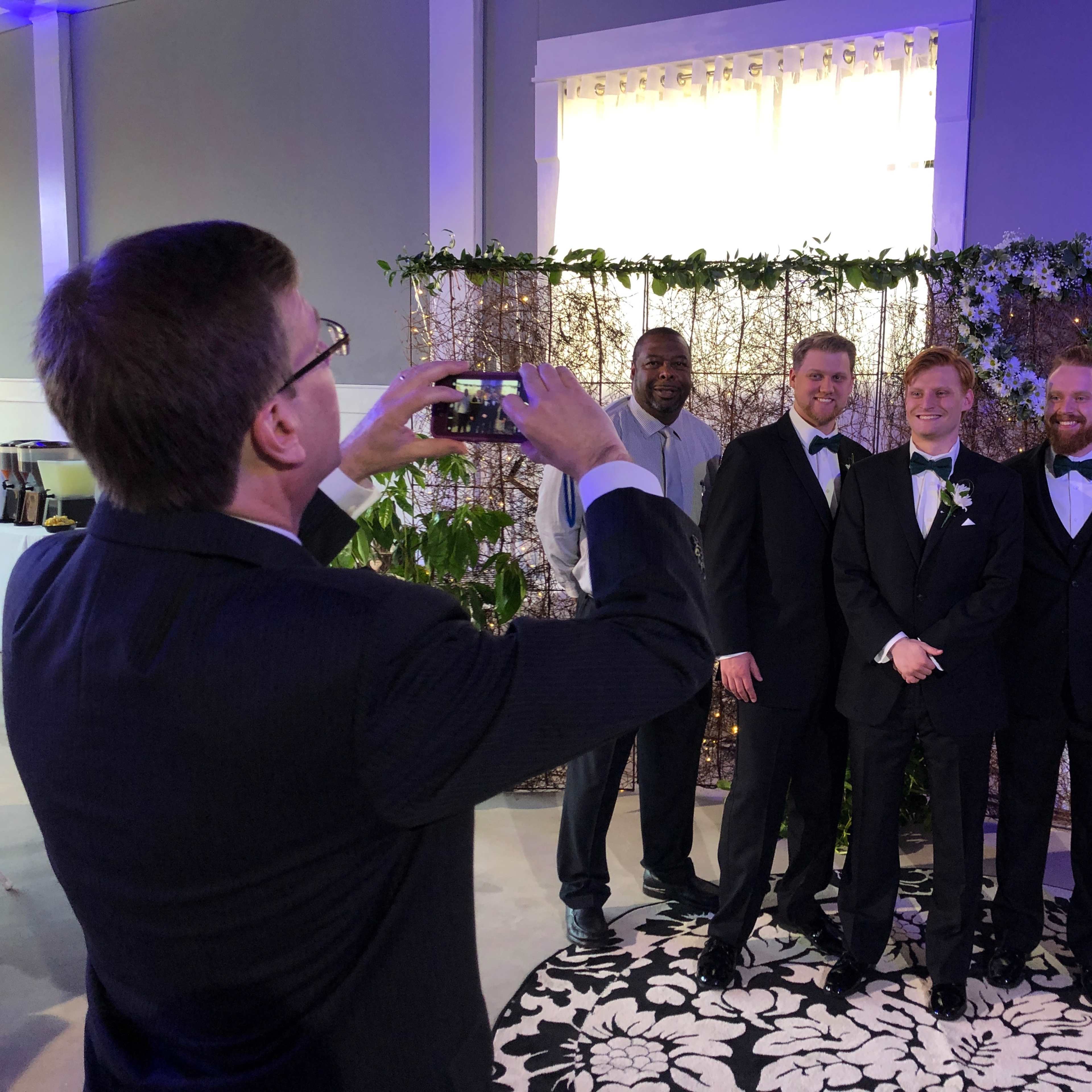 A group of five people, dressed in formal attire, pose for a photo in front of a floral backdrop at a wedding reception.
