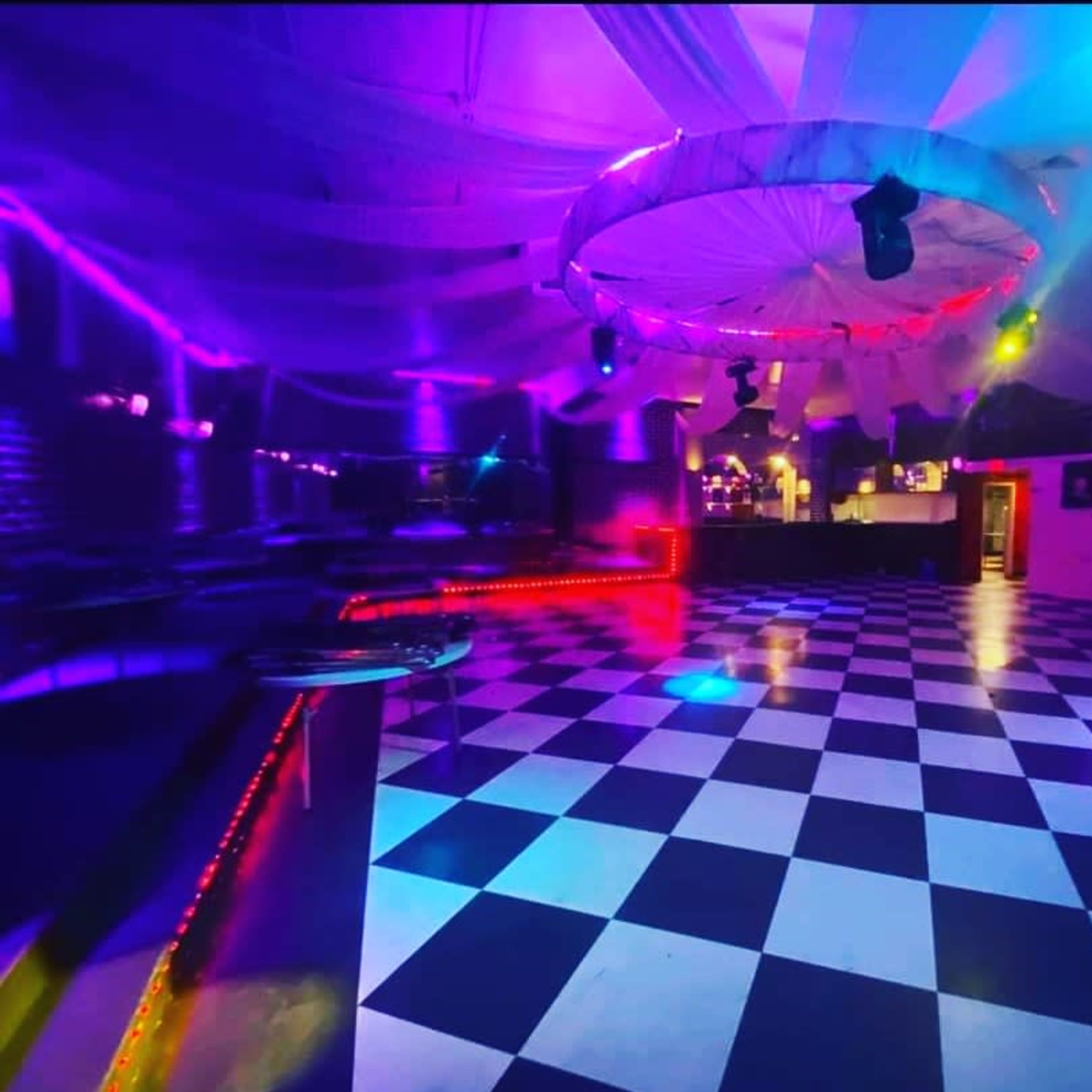 An empty dance floor with a black and white checkered pattern, illuminated by colorful lights and draped fabric on the ceiling.