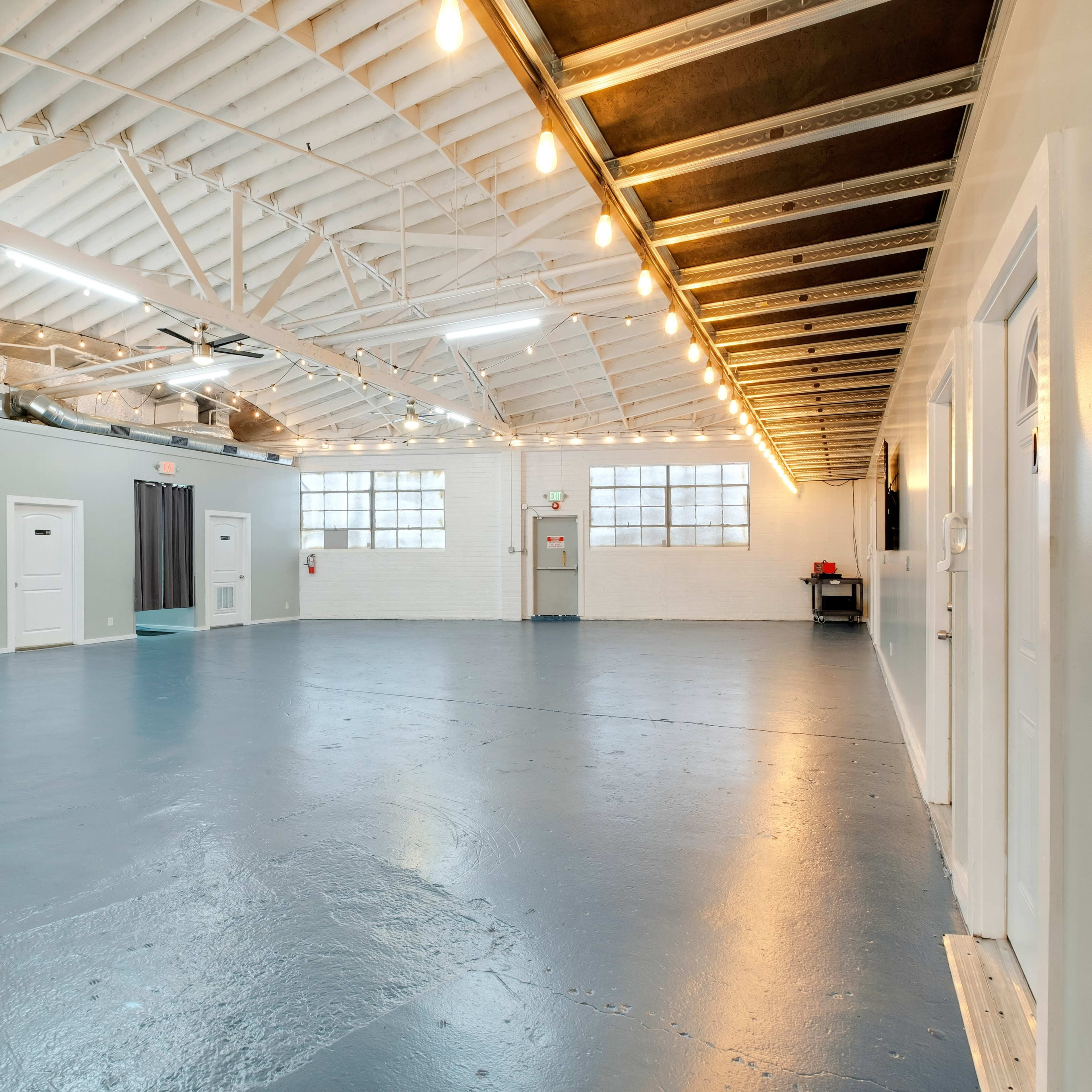 A spacious, well-lit industrial room featuring a polished blue floor, exposed ceiling beams, and several white doors along the walls.