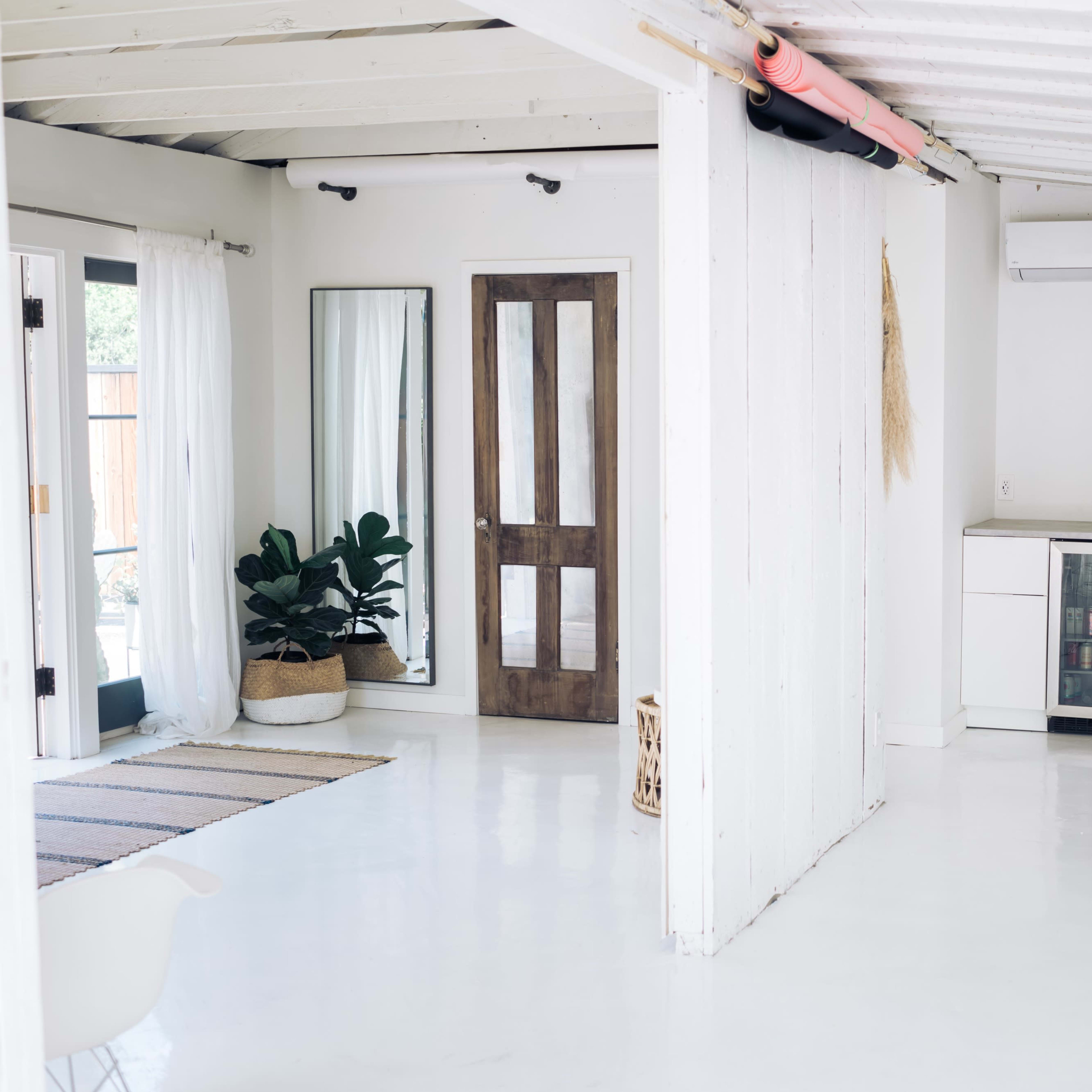 A bright, minimalist interior space featuring a white floor, a wooden door, a large mirror, a small fridge, and a potted plant.