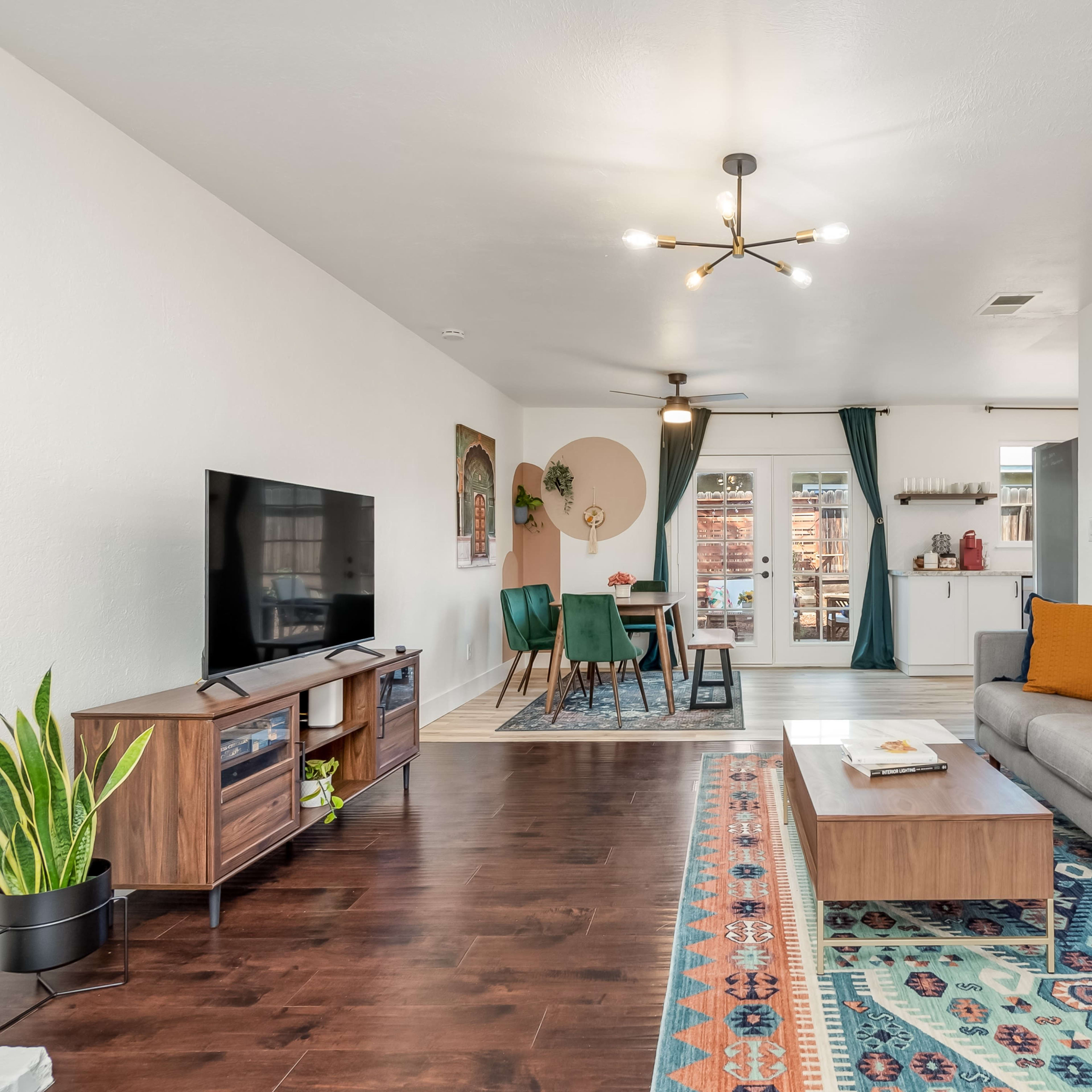 The living room features a gray sofa, a wooden coffee table, a TV stand with a television, and a dining area in the background, all accented by a colorful area rug and large windows.