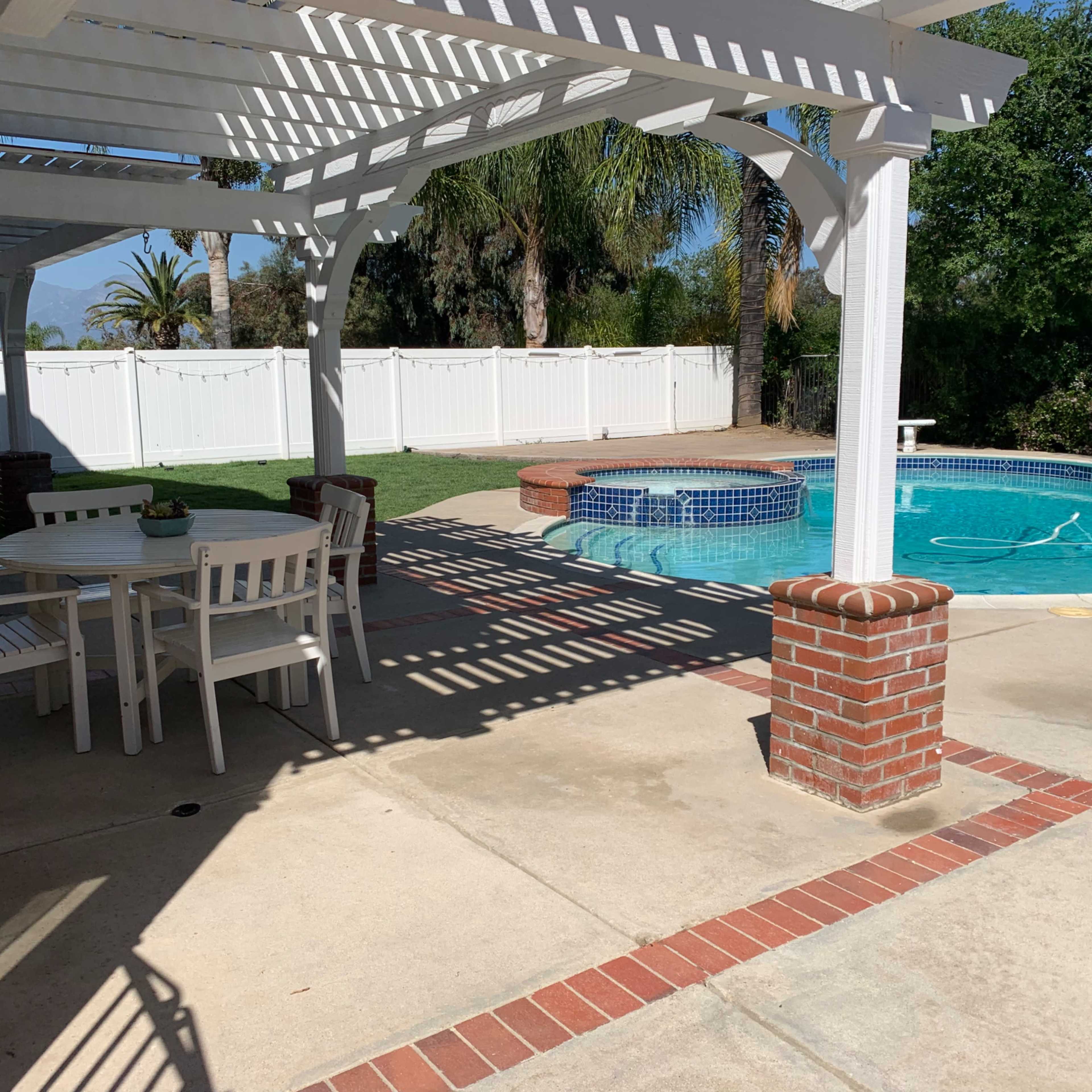 A backyard with a swimming pool surrounded by a patio, a white pergola, and a dining table set for four.