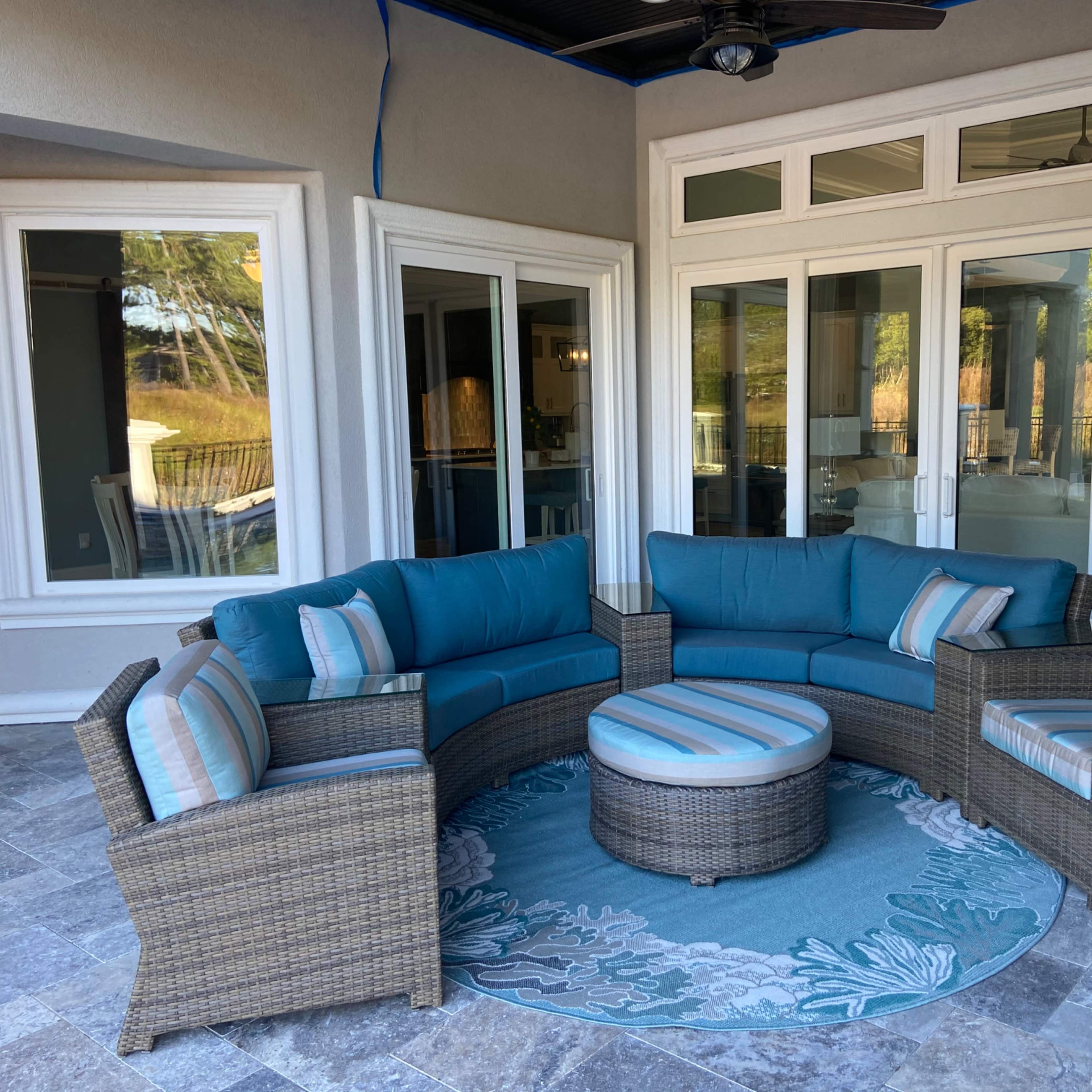 A patio area featuring a circular seating arrangement in blue cushions, a matching round coffee table, and a decorative rug on a tiled floor, enclosed by large glass doors.