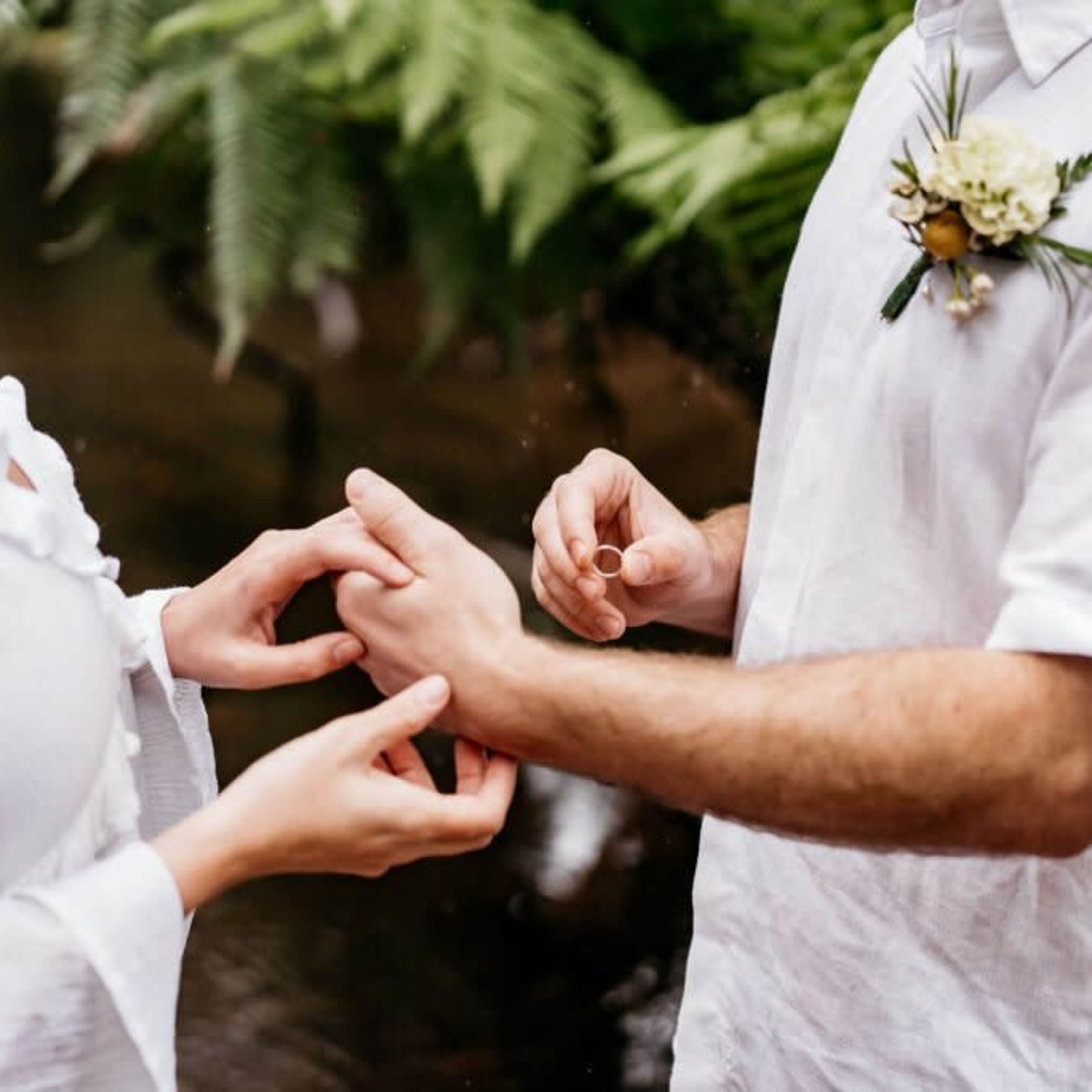 A person wearing a white shirt places a ring on another person's finger in a lush outdoor setting.