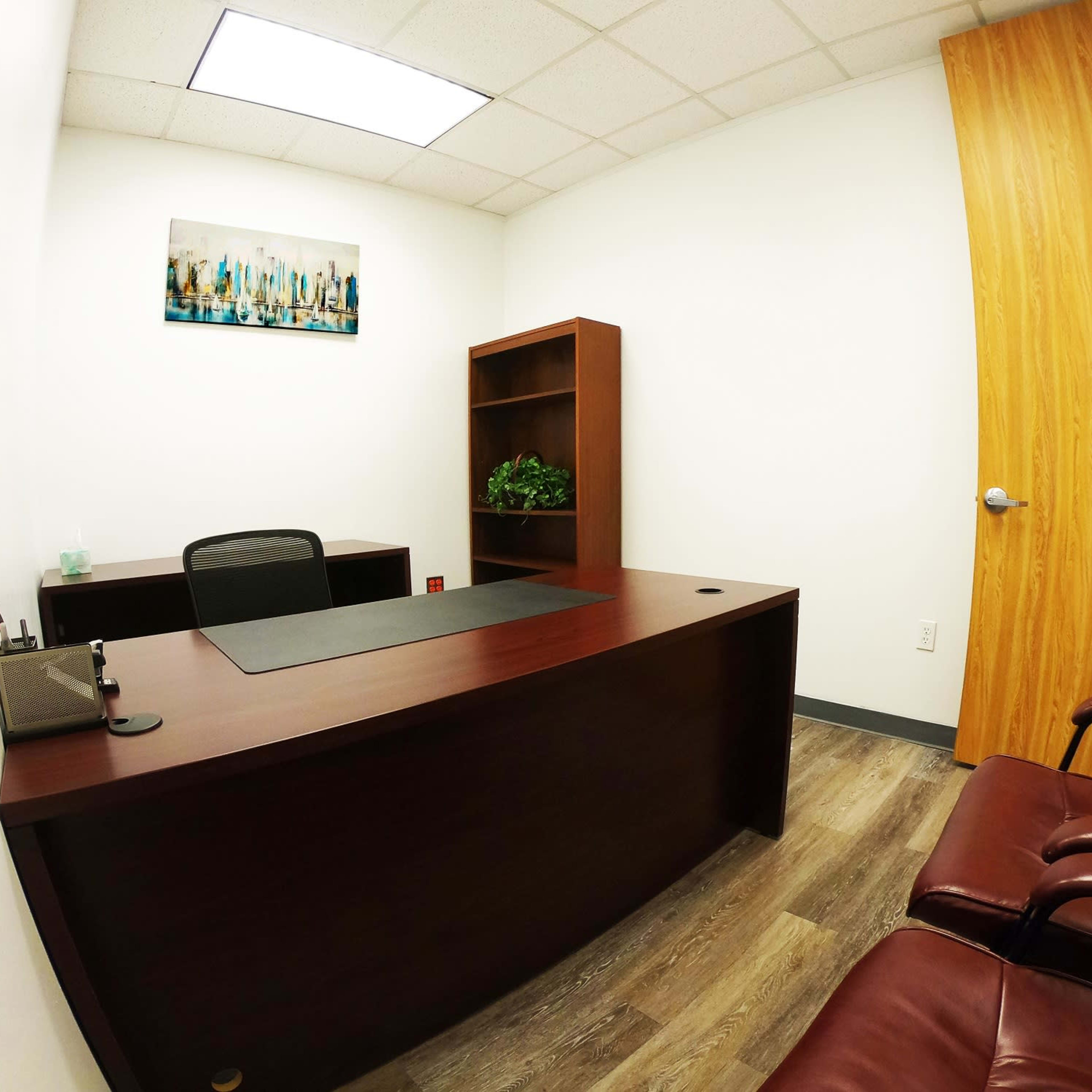 A small office with a wooden desk, a chair, a bookshelf, and a door leading outside.