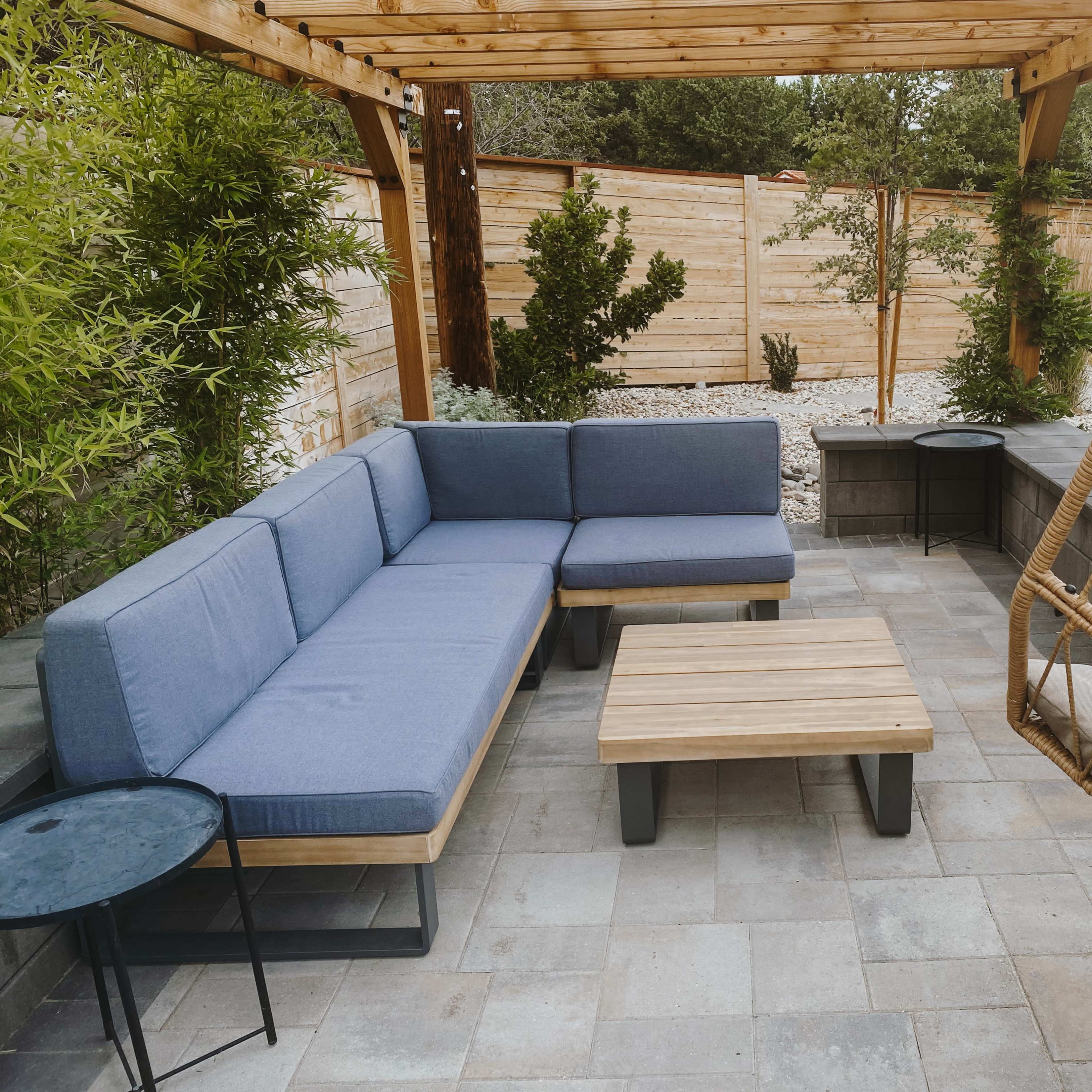 A cozy outdoor seating area featuring a blue sectional couch, a wooden coffee table, and a hanging chair, all surrounded by greenery and stone pathways.