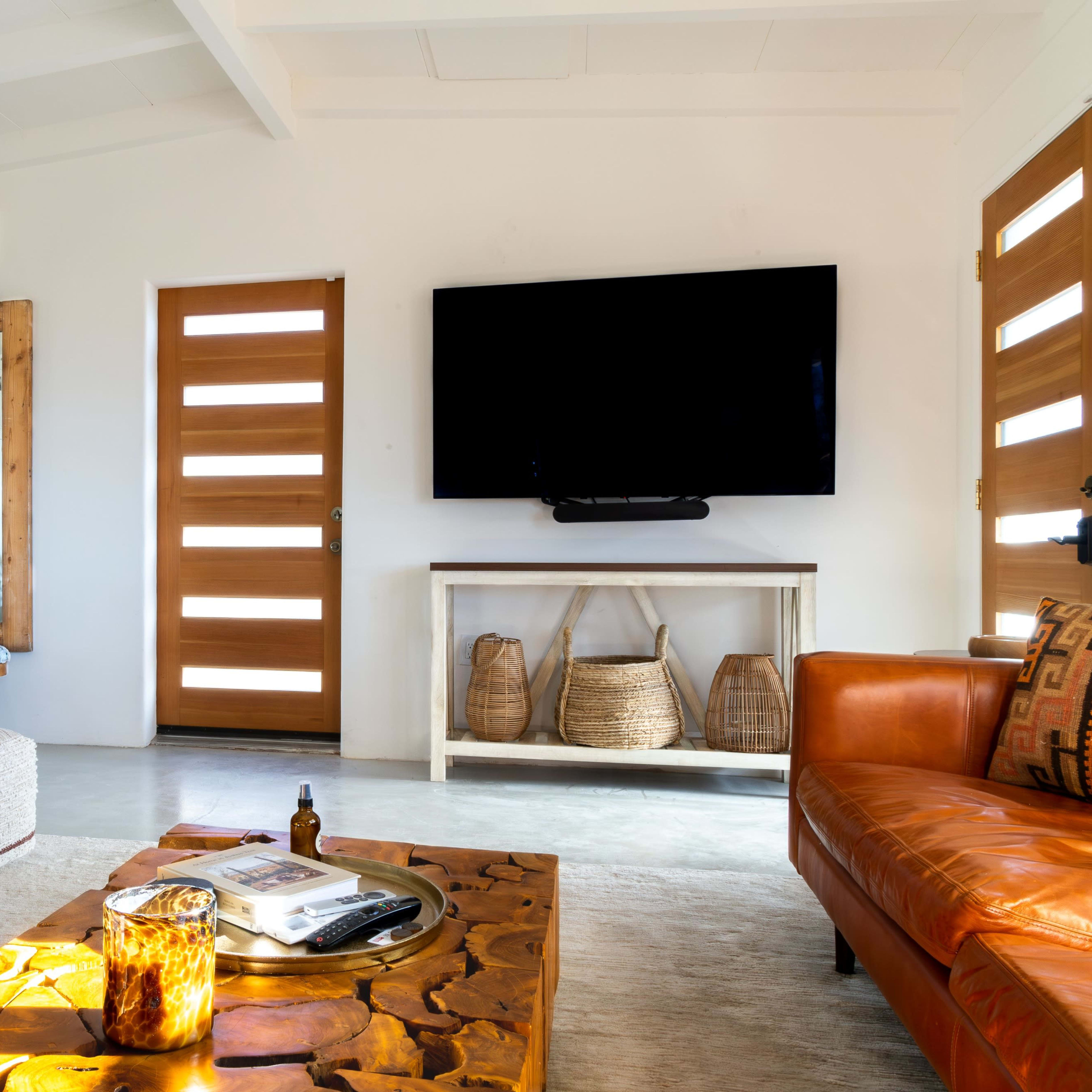 The living room features a leather sofa, a wooden coffee table, and a television mounted on the wall, with a mirror and wooden doors in the background.
