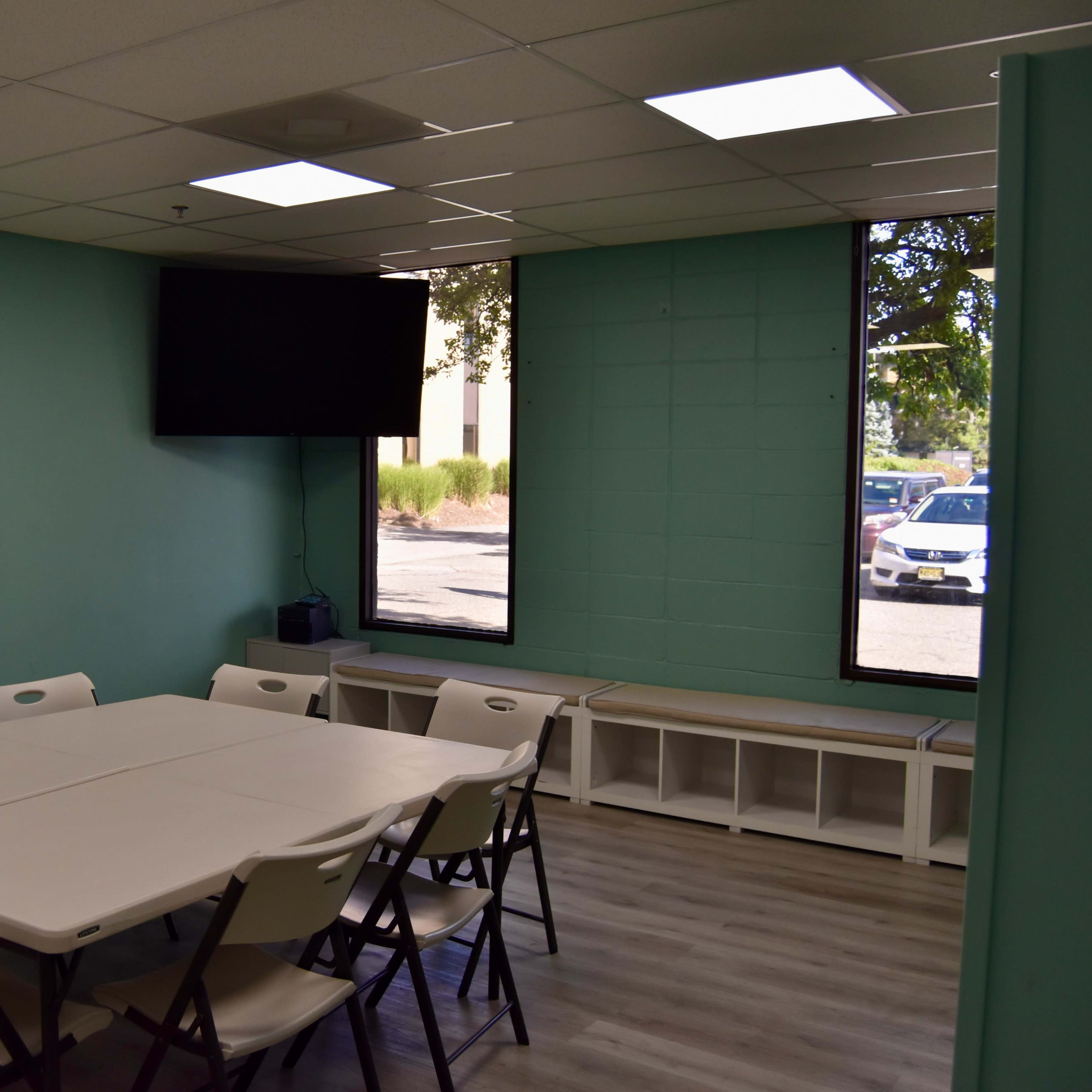 A meeting room features a rectangular table surrounded by eight folding chairs, with a mounted television on the wall and large windows allowing natural light.