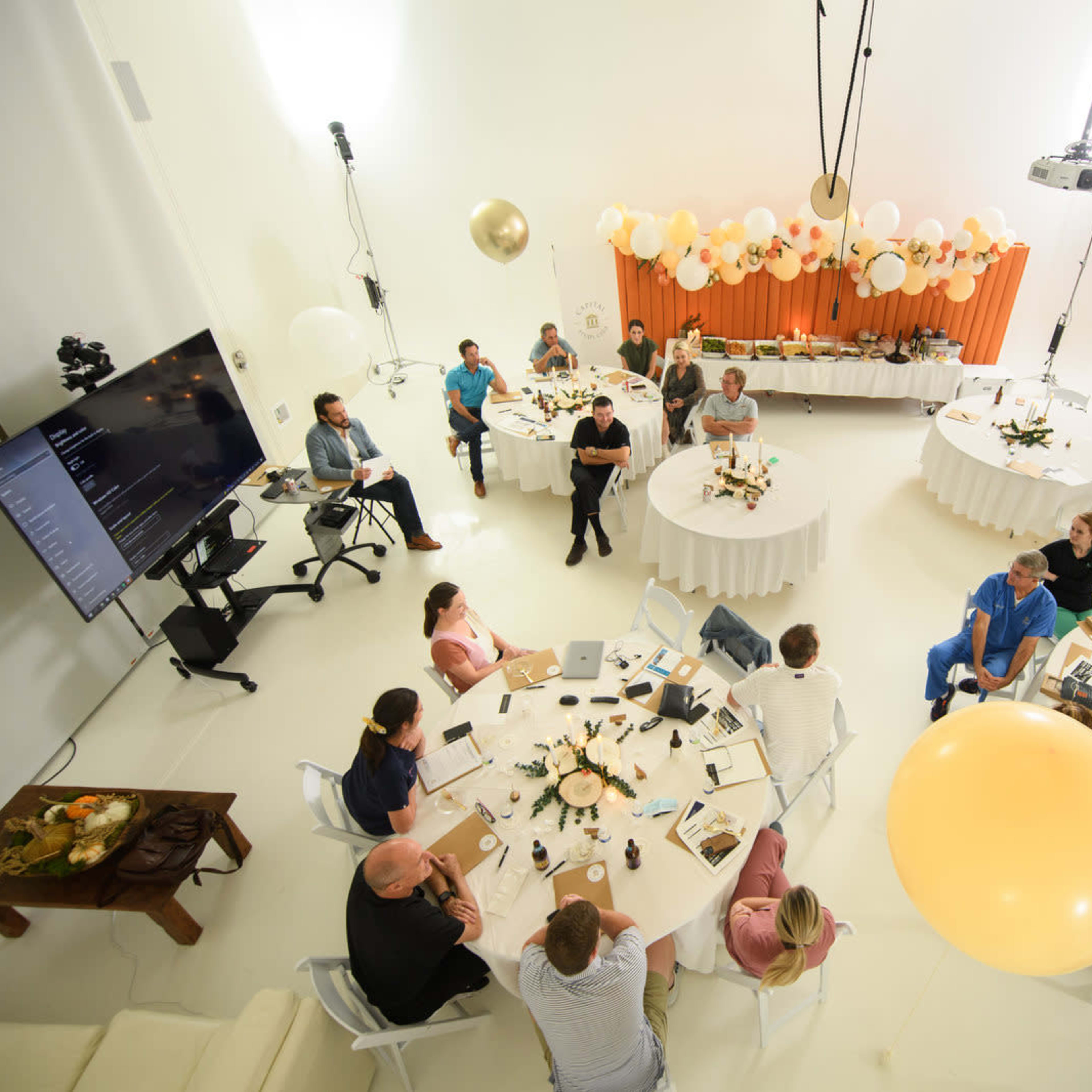 A round table meeting is taking place in a bright, minimalistic room with decorations including balloons and floral arrangements.