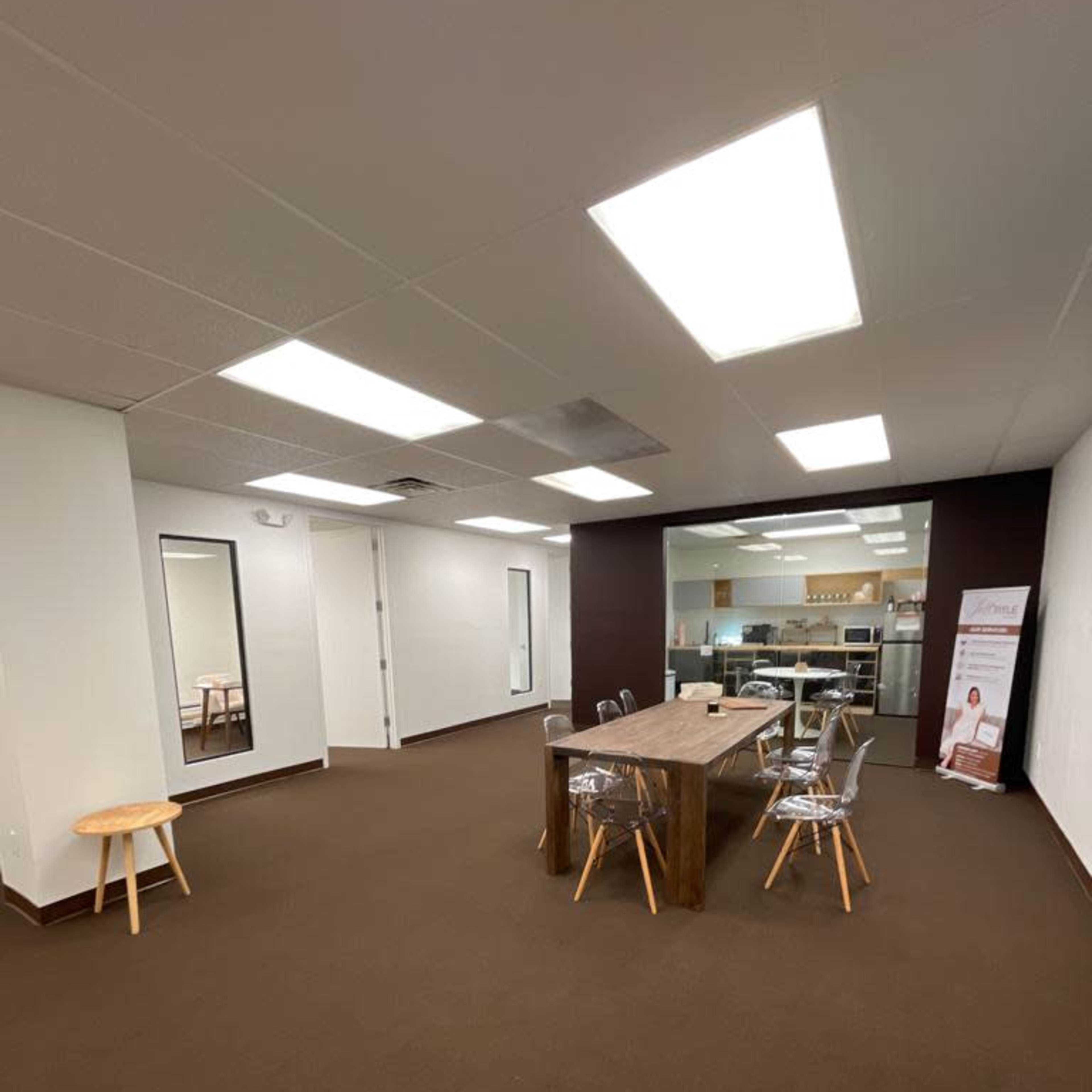 A minimalist office space featuring a large wooden table surrounded by clear chairs, with a mirror and a kitchenette visible in the background.