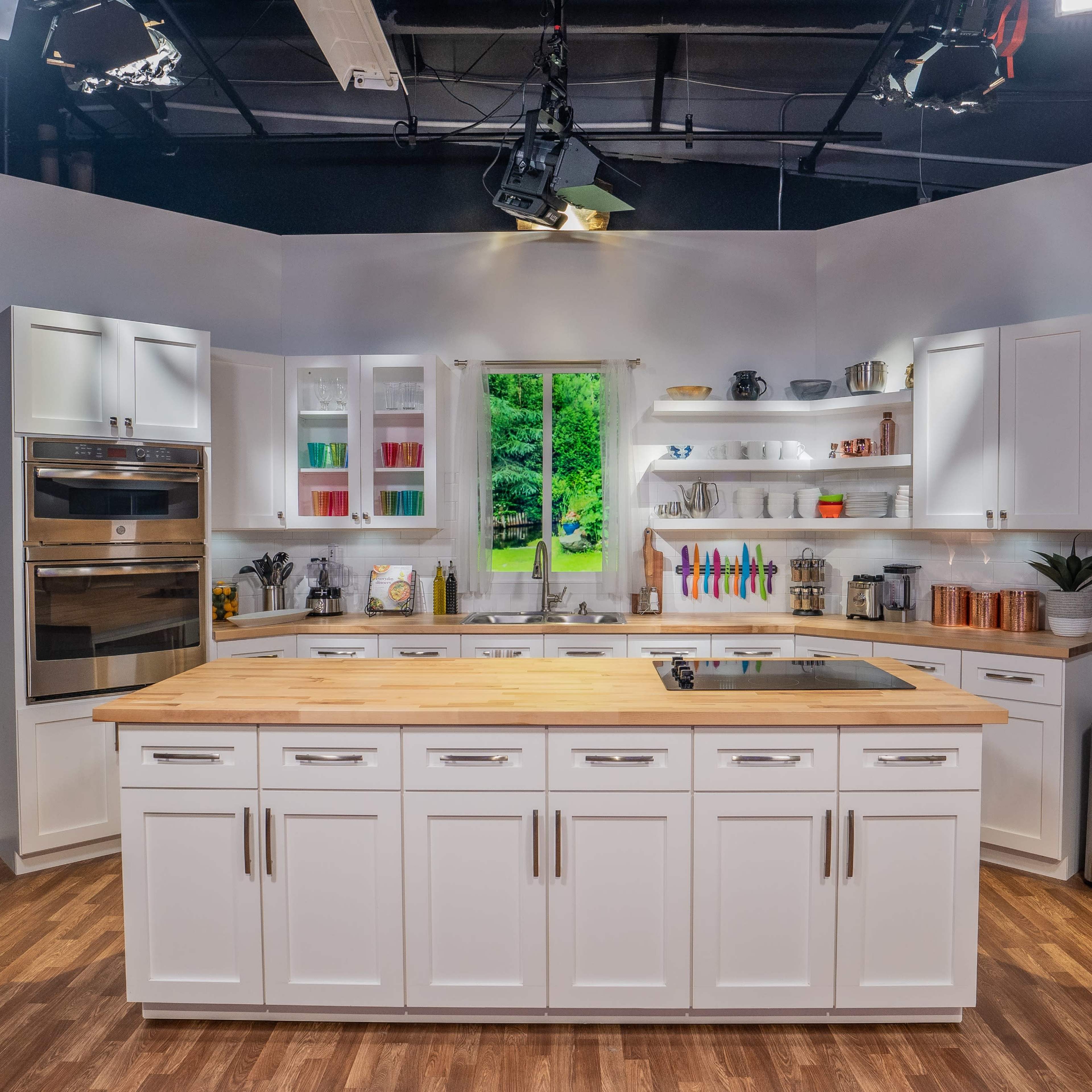 A modern kitchen set features a central island with a wooden countertop, surrounded by white cabinetry and shelves filled with kitchenware, set against a backdrop of a large window offering a view of greenery.