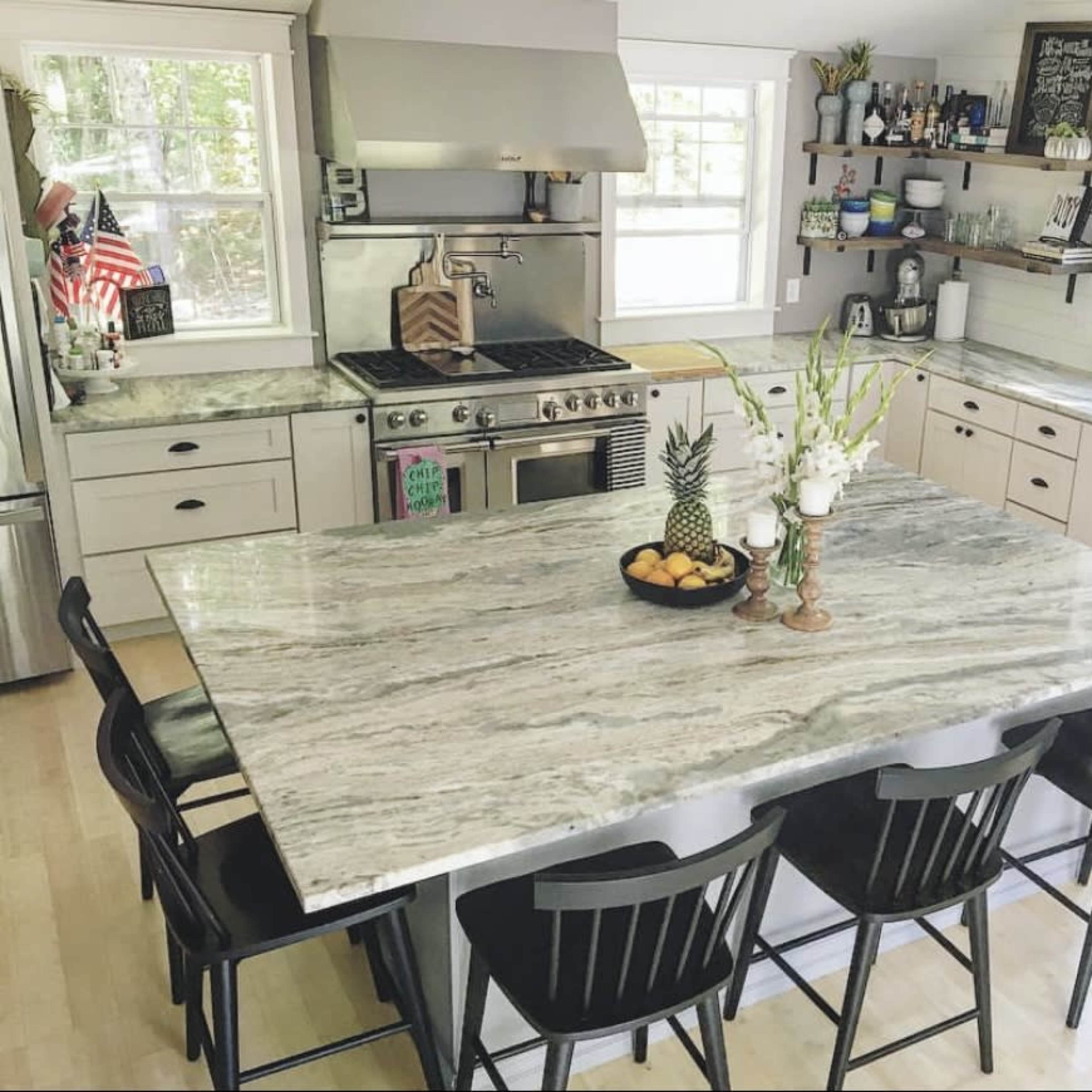 The kitchen features a large island with a granite countertop, surrounded by black chairs, and includes stainless steel appliances and shelves displaying various kitchen items.
