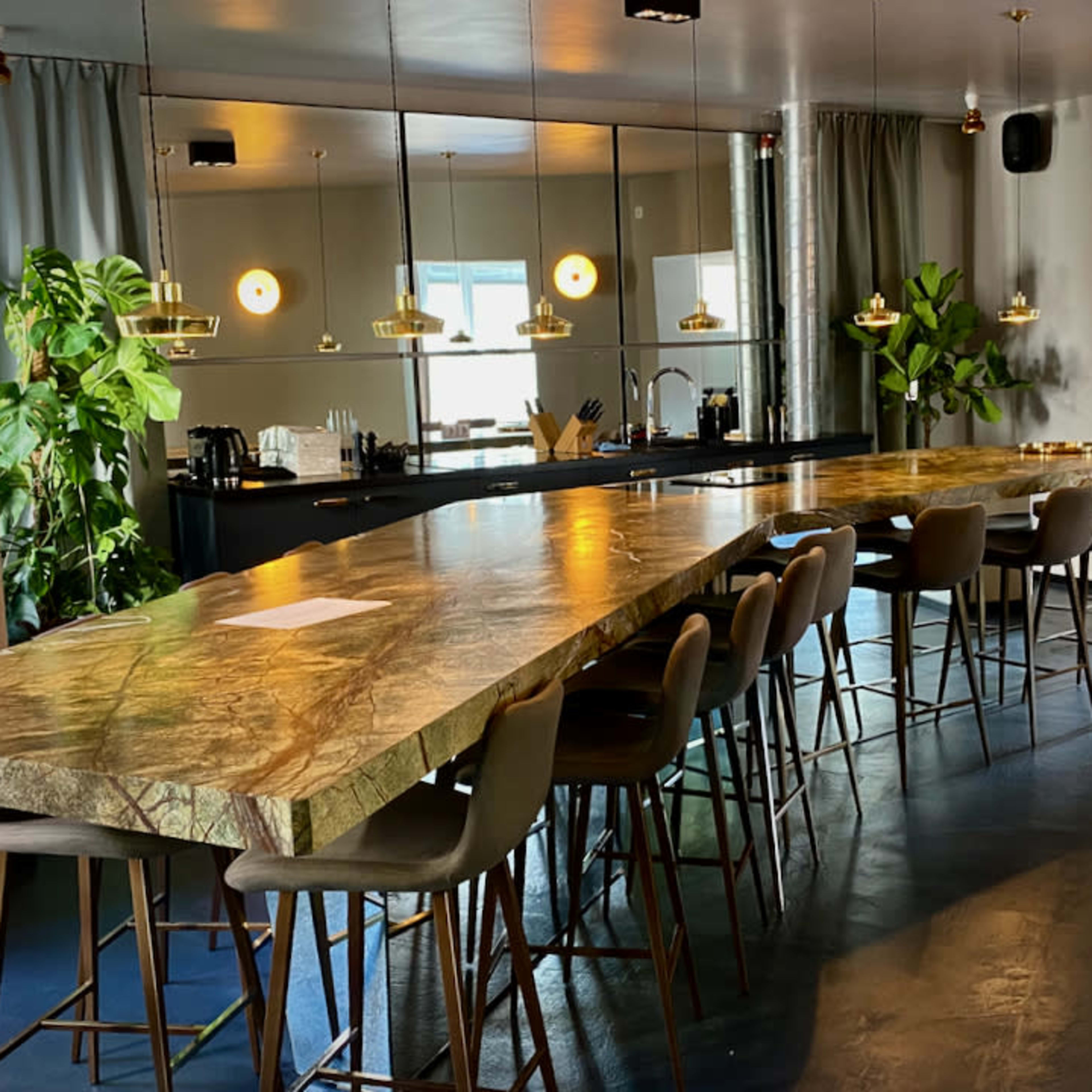 A long marble countertop with high stools is featured in a modern, well-lit kitchen area with plants and large mirrors.