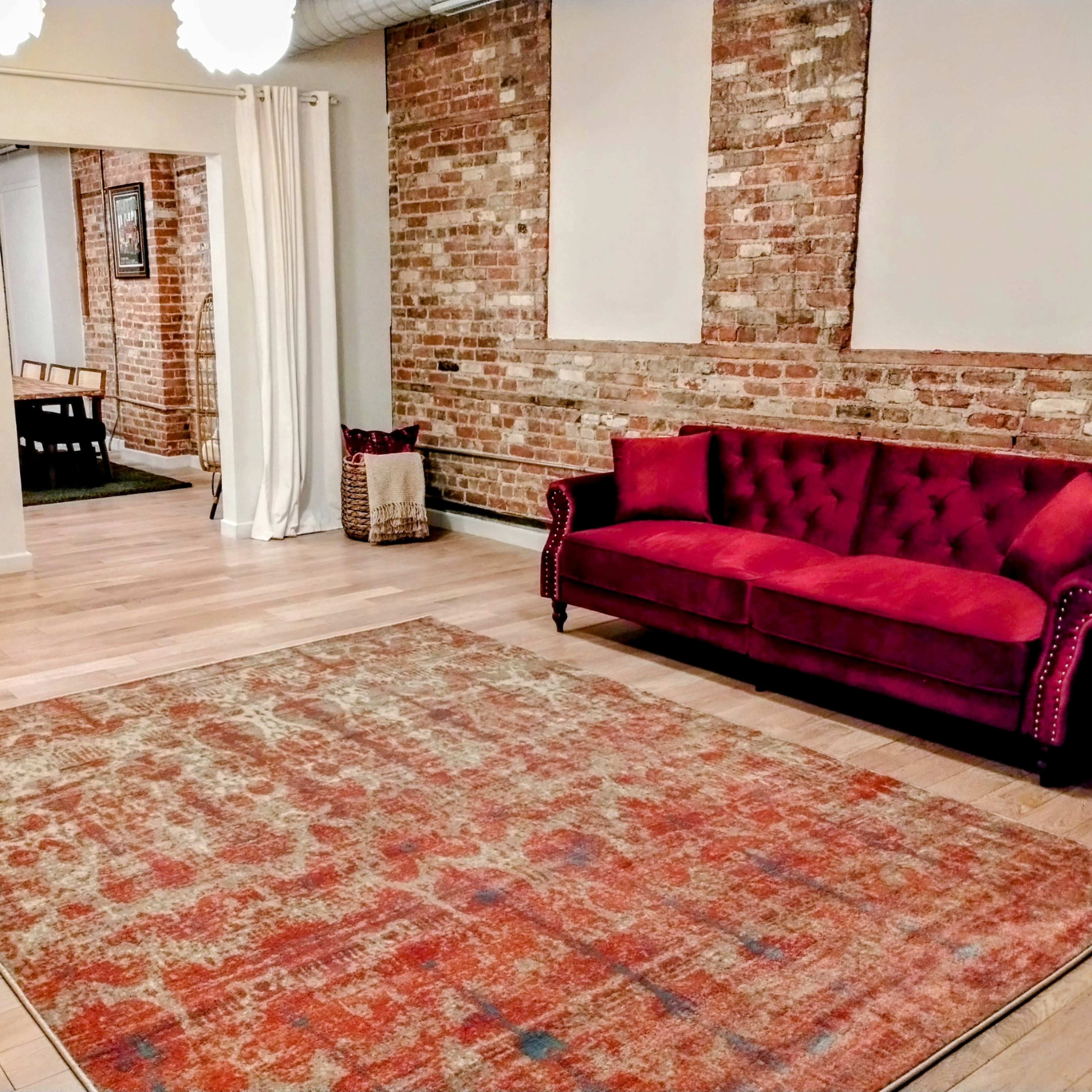 A red couch is positioned on a patterned area rug in a room with exposed brick walls and light wooden flooring.