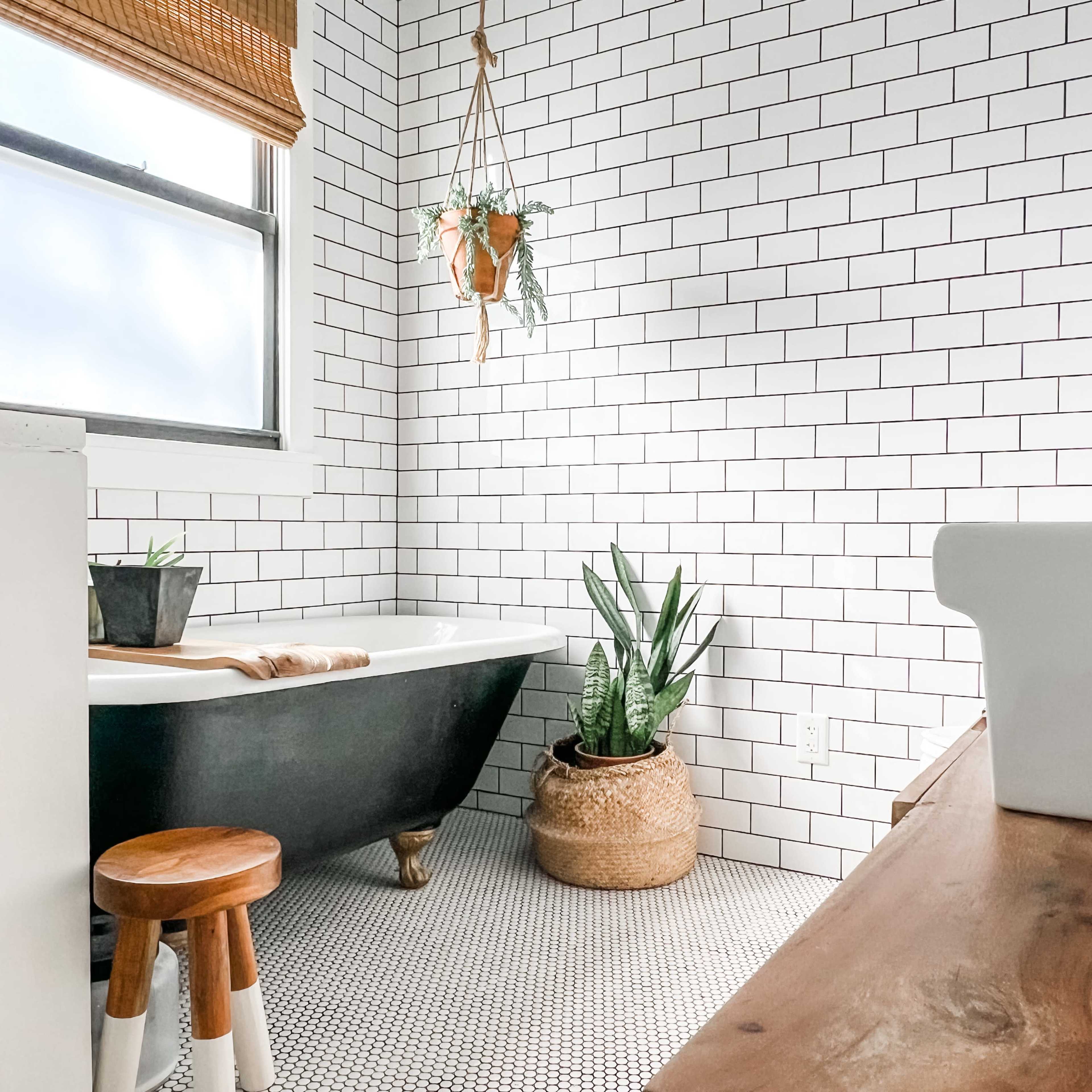 A bathroom featuring a black freestanding tub, a wooden stool, potted plants, and white subway tile walls.