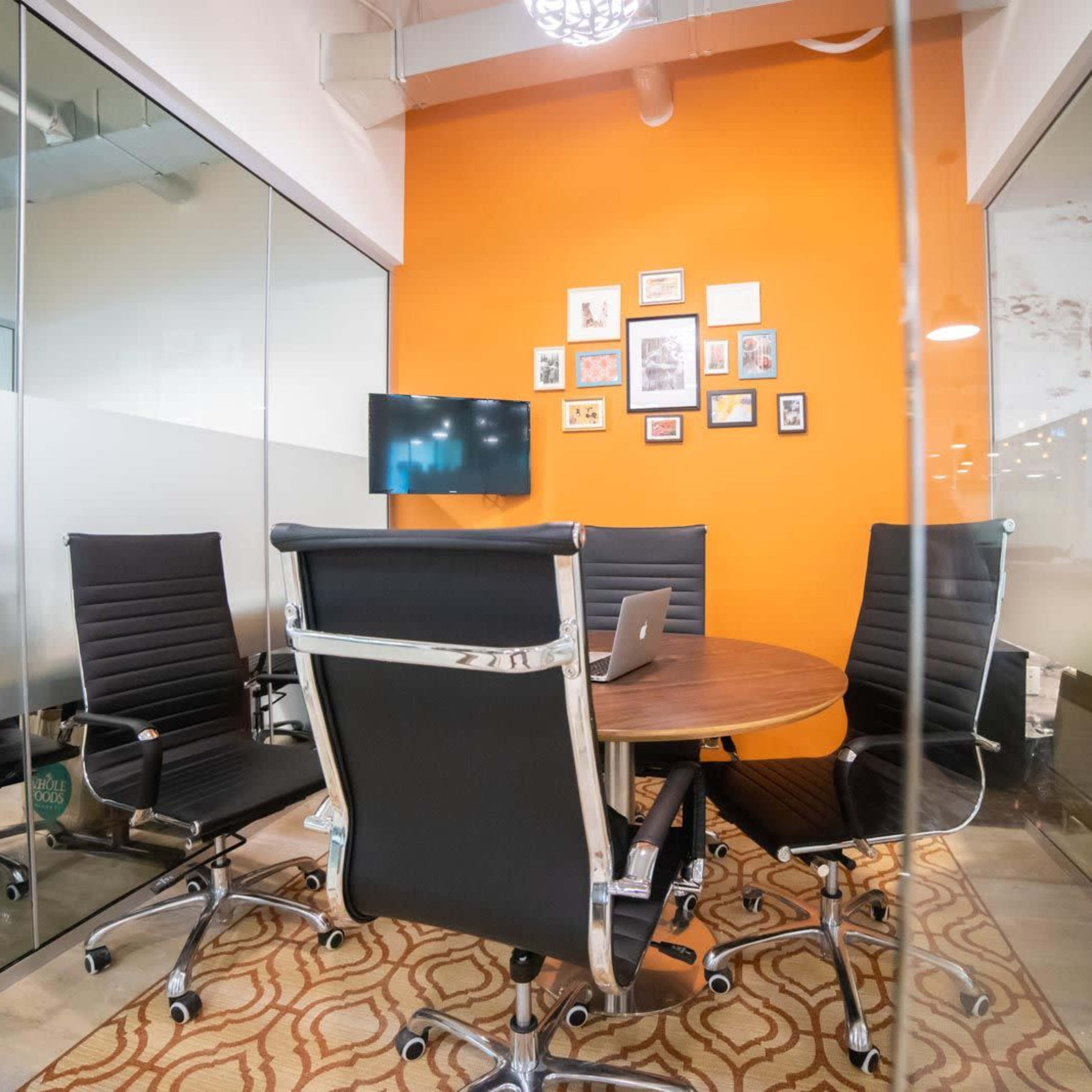 A modern conference room features a round table surrounded by black chairs, with an orange wall displaying framed pictures and a mounted TV.