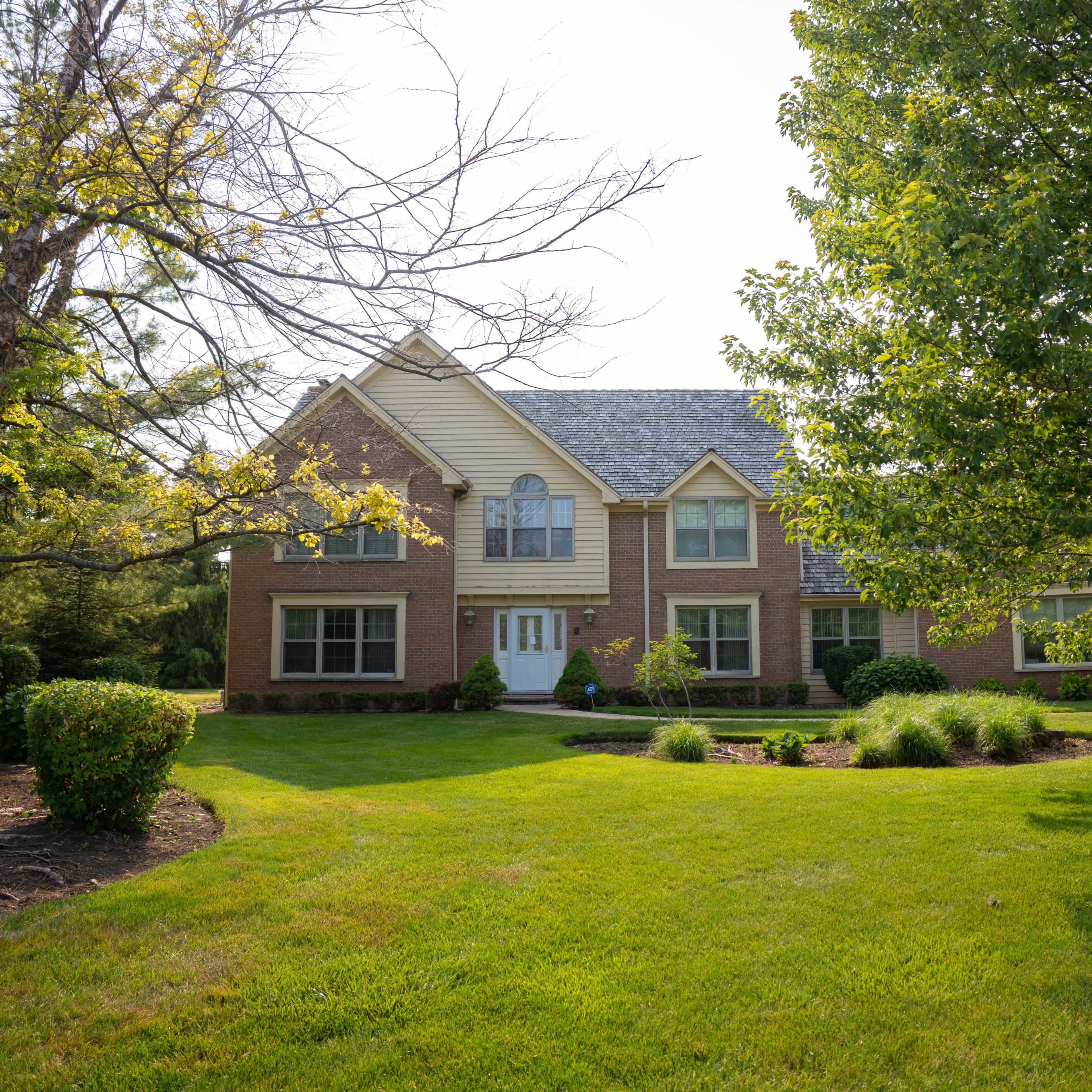 A brick house with a pitched roof is set on a landscaped lawn surrounded by trees and shrubs.