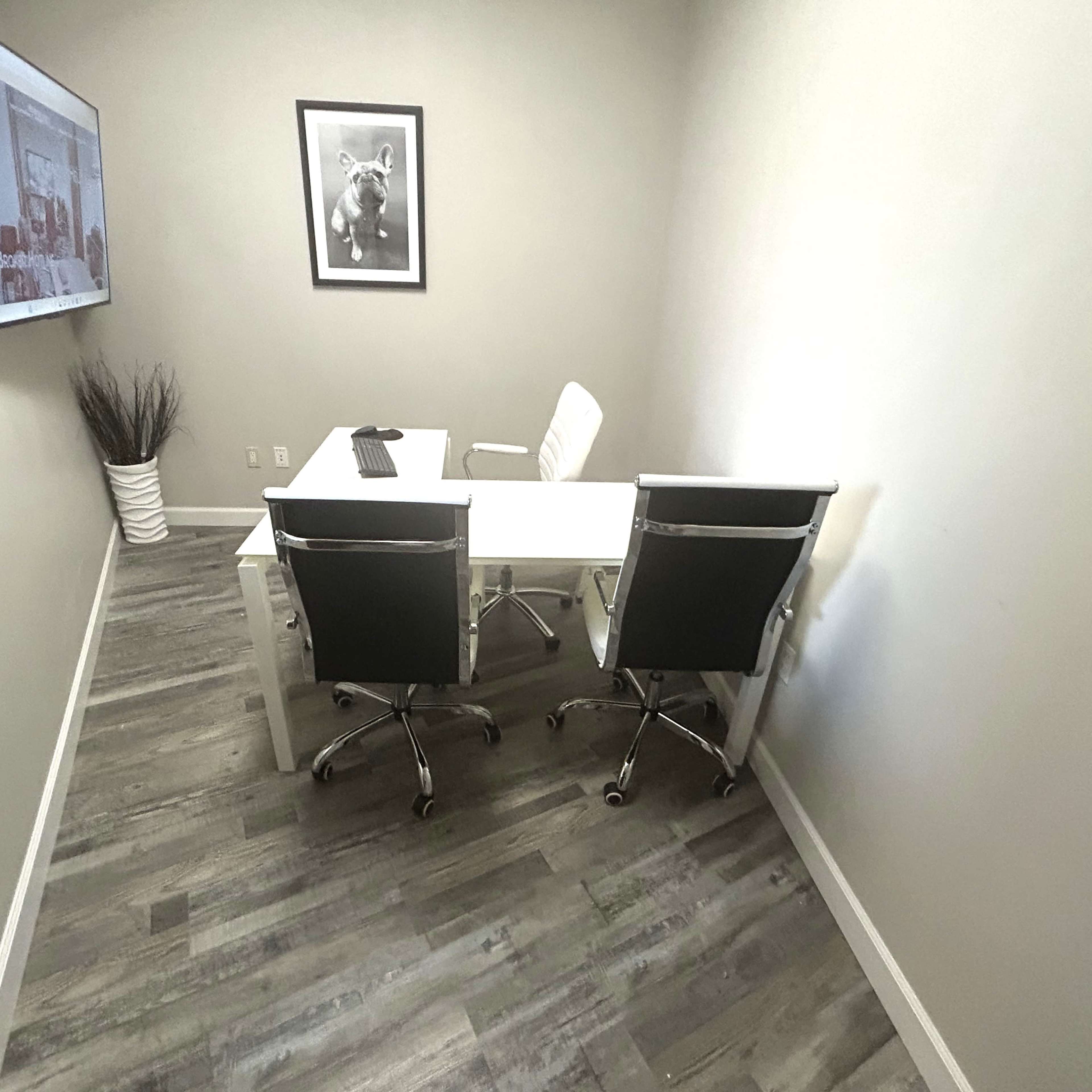 A modern, small office space features a white desk with two chairs, a wall-mounted screen, and a framed picture.