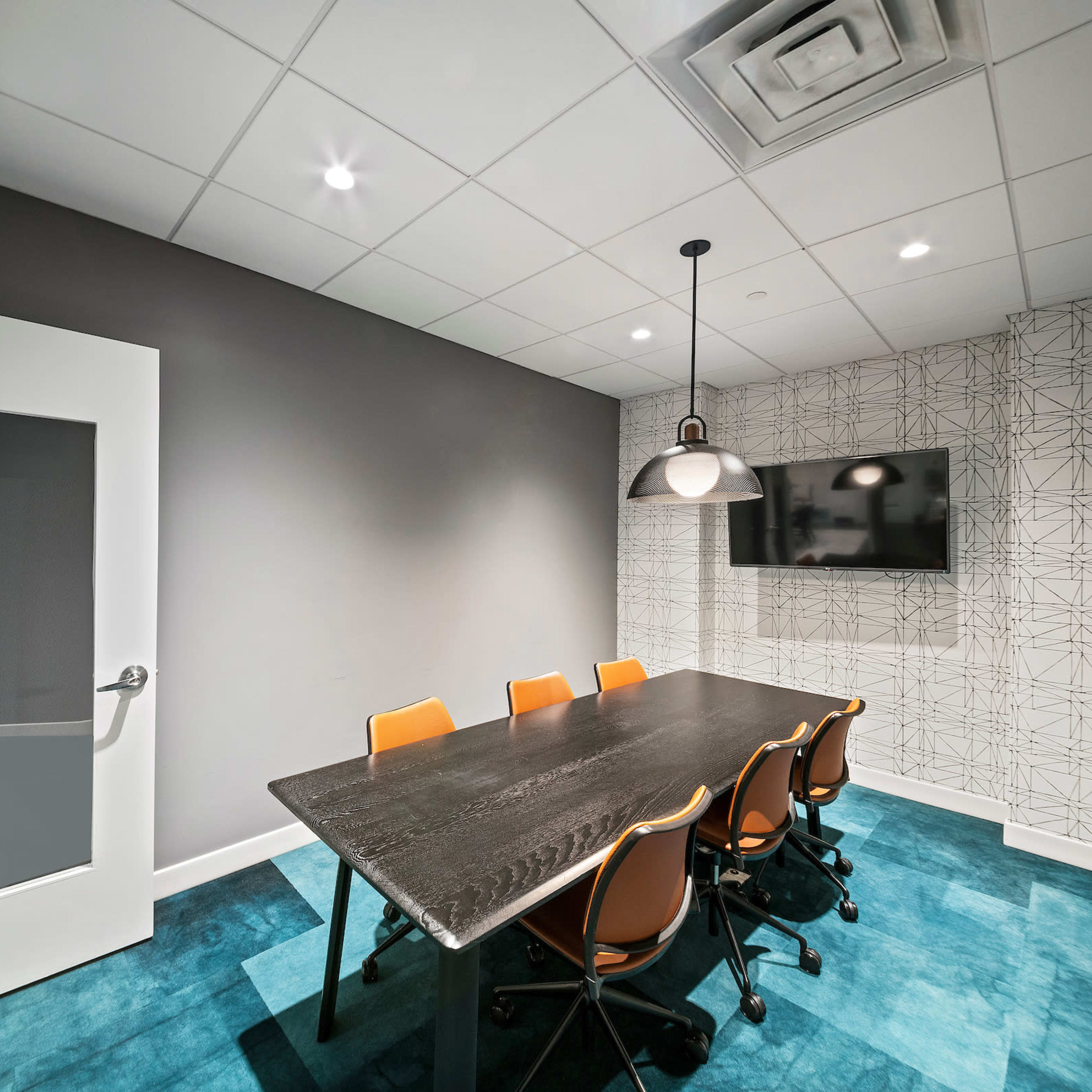 A modern conference room features a black wooden table surrounded by four orange chairs, a wall-mounted TV, and a patterned accent wall.