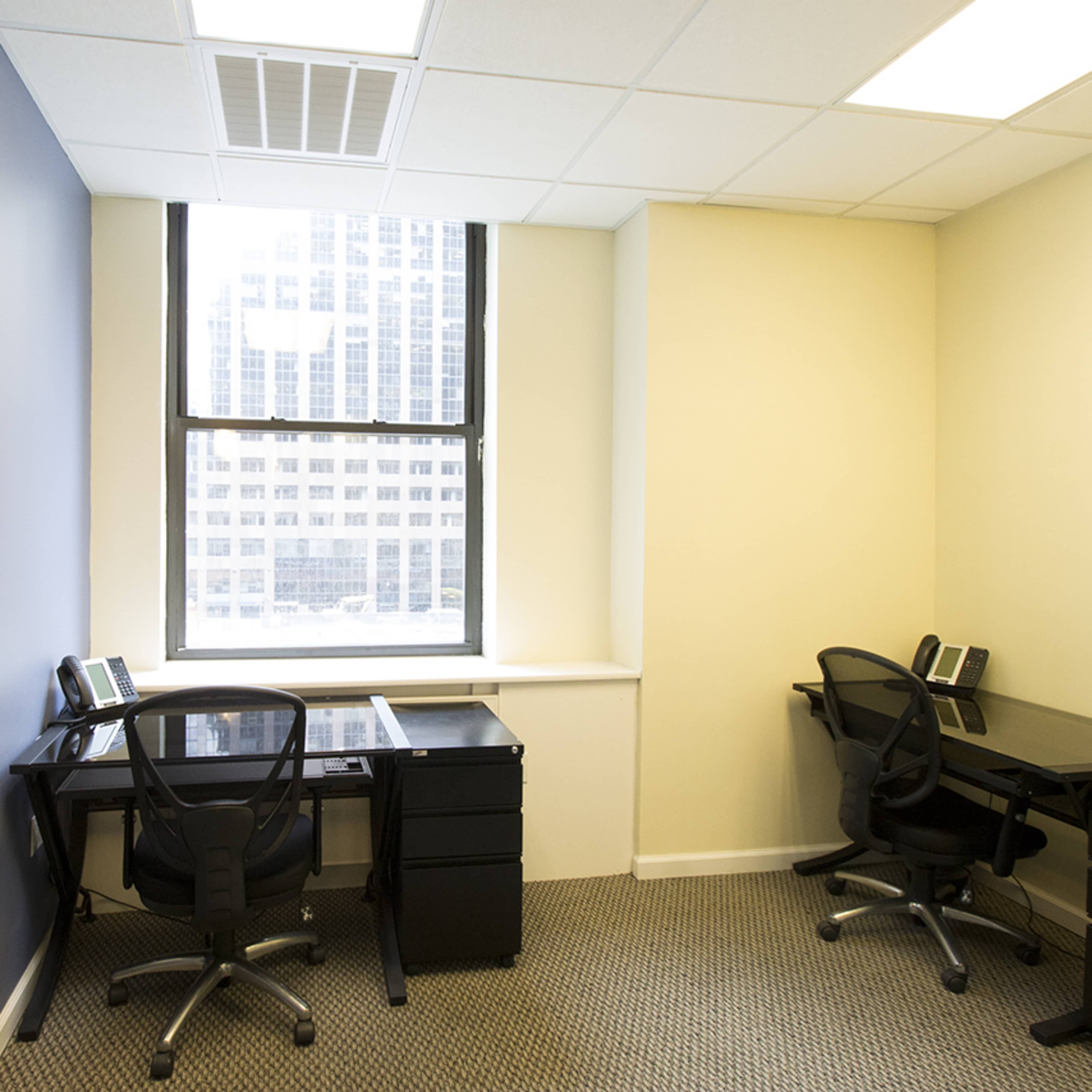 A small office space with two black desks and chairs, separated by a window overlooking a cityscape.