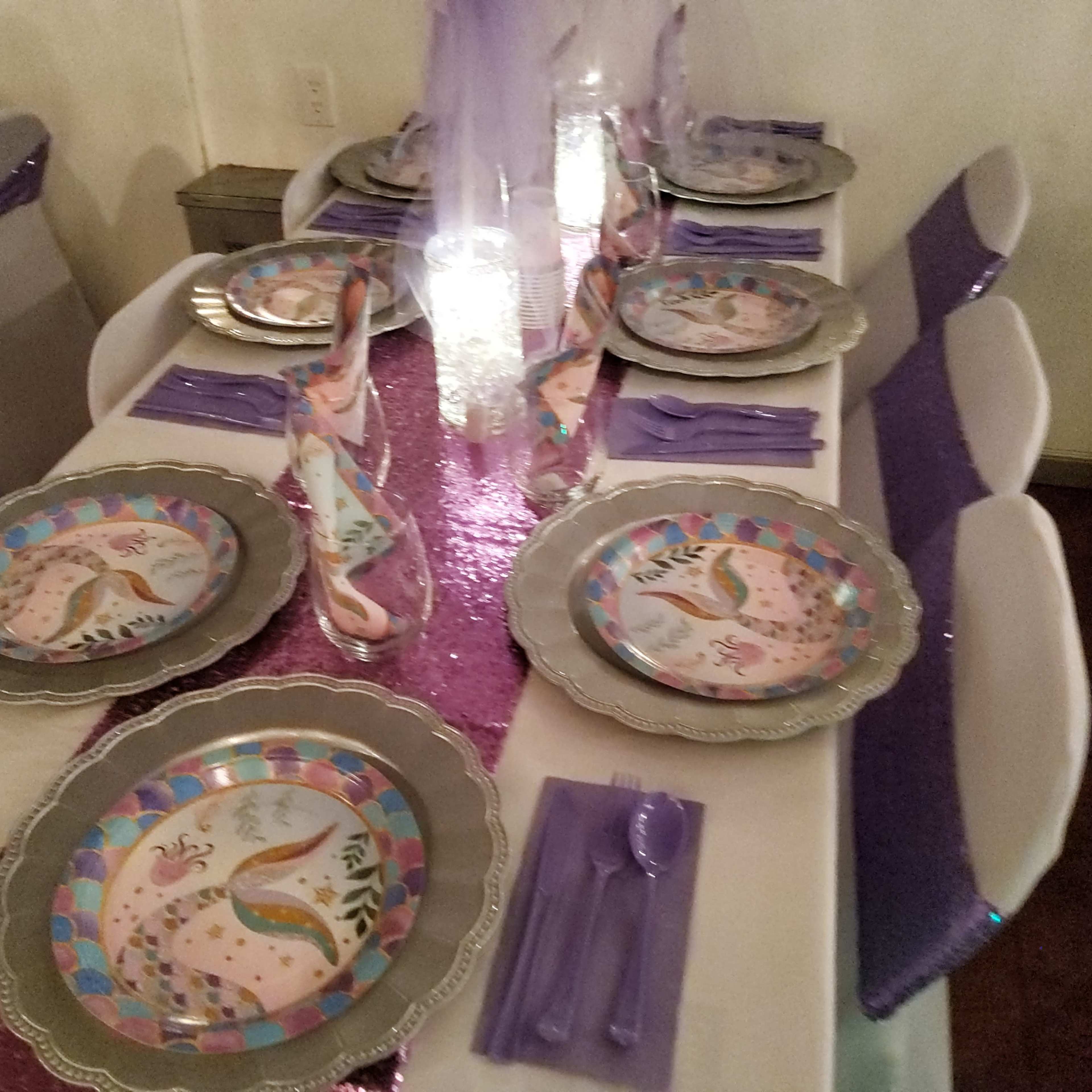 A decorated dining table is set with plates, utensils, and purple-themed decorations, featuring a sparkling table runner and colorful napkins.