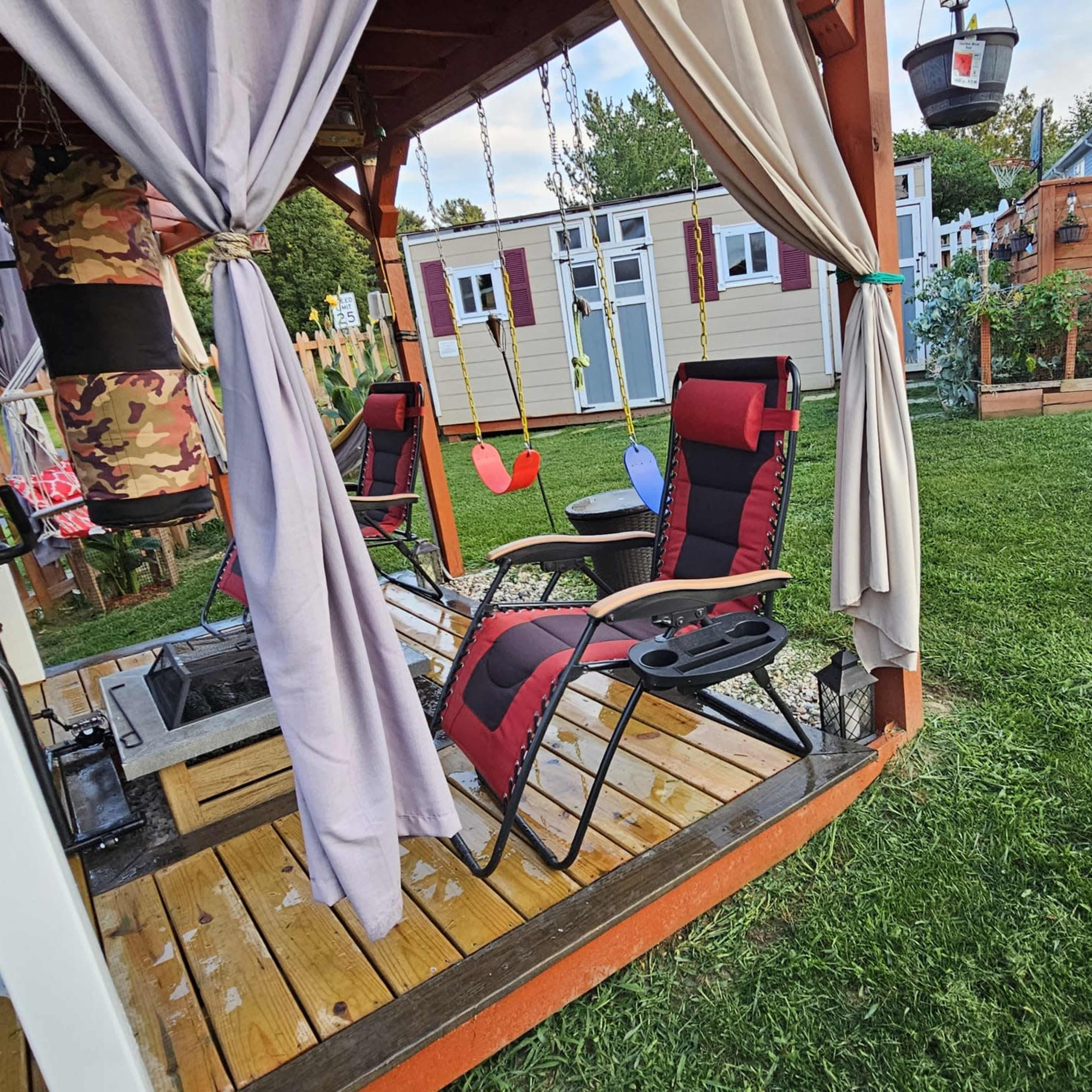 A gazebo with two reclining chairs and a small table is situated on a wooden deck in a backyard, surrounded by green grass and a vegetable garden.