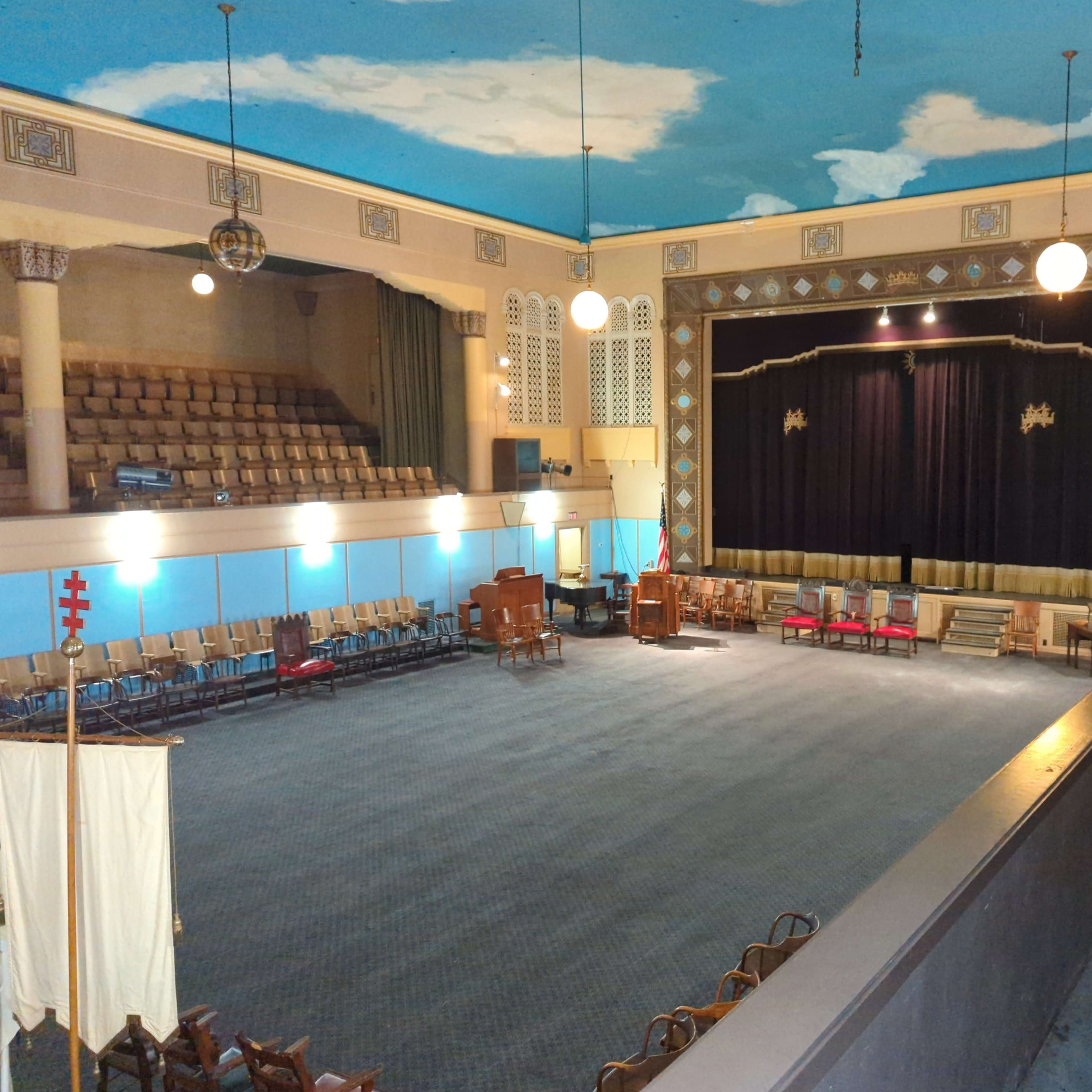 A spacious auditorium with a dark stage, featuring rows of empty chairs and a ceiling painted with clouds.