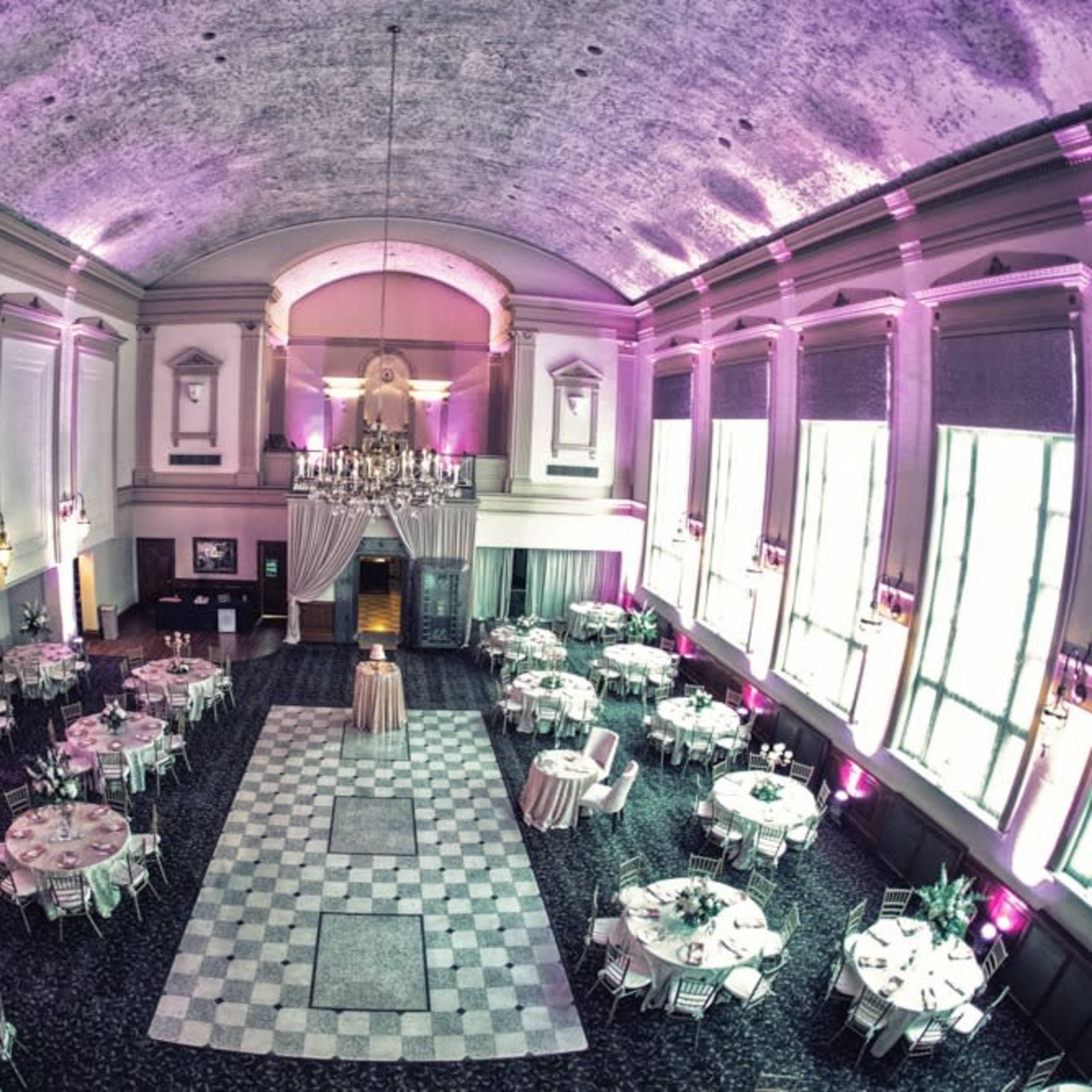 An elegantly arranged banquet hall with round tables, a checkered dance floor, and purple lighting illuminating the high ceiling.