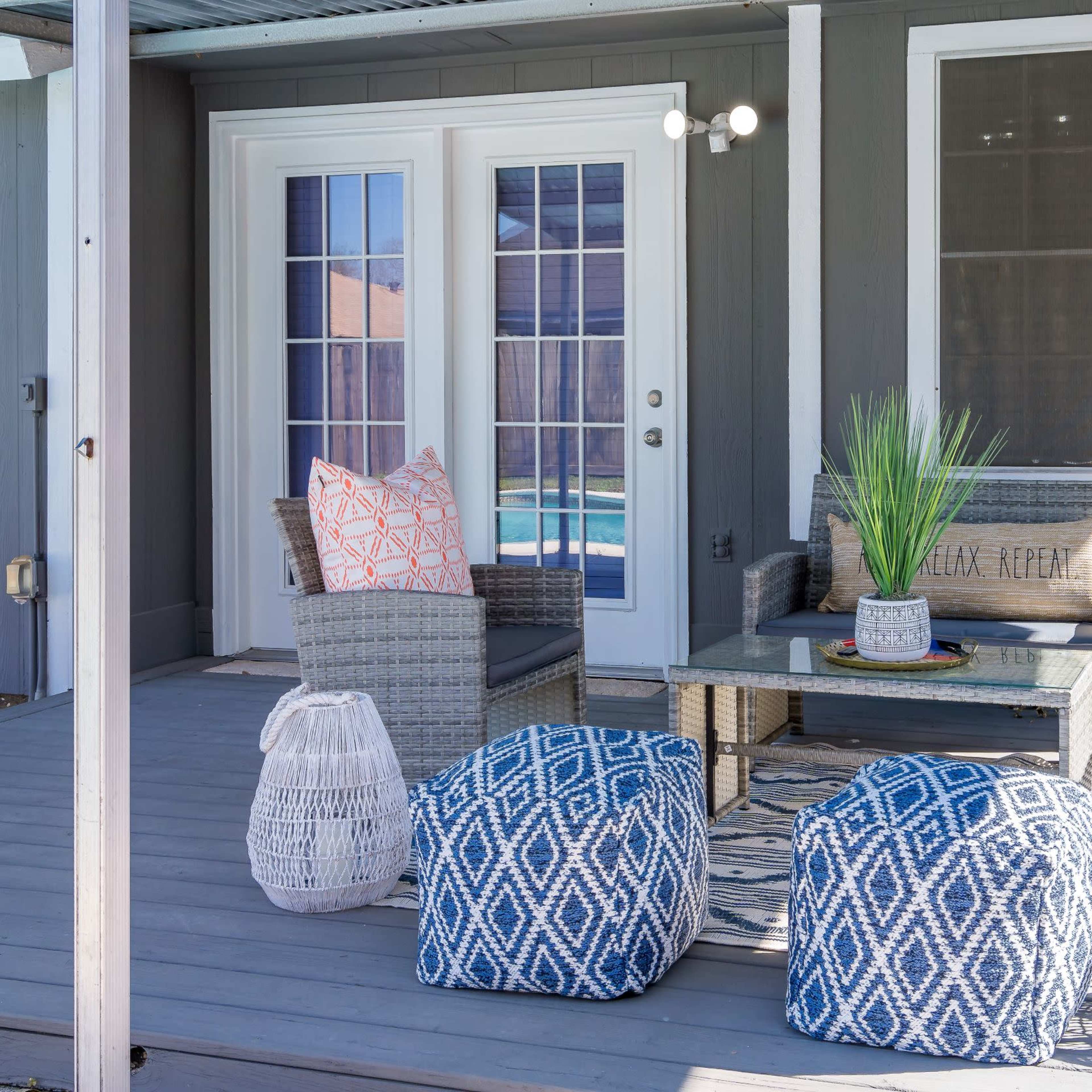A patio area with two woven chairs, a small table, and decorative cushions, surrounded by poufs and a patterned rug.