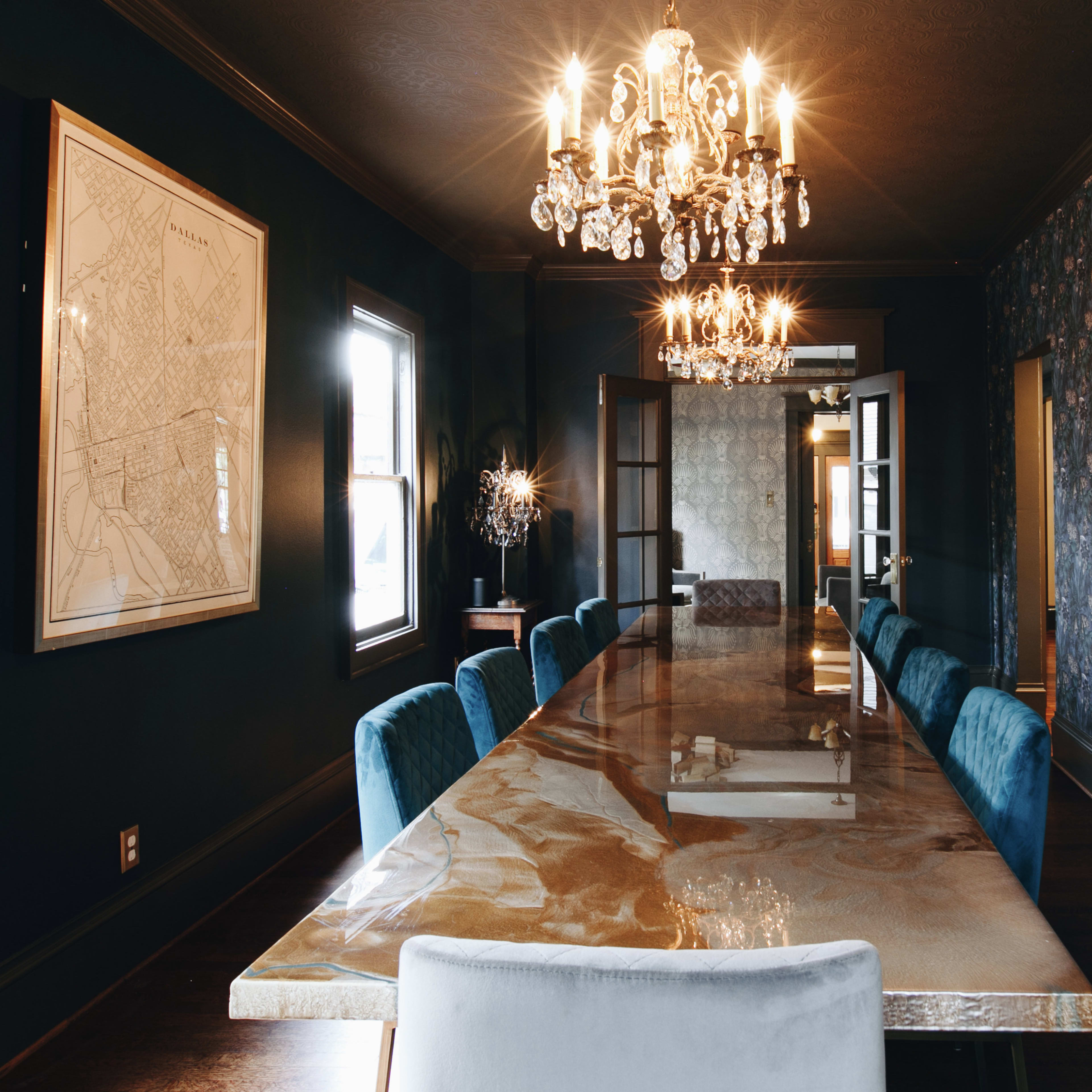 A long table with a glossy surface and blue chairs is centrally positioned in a dimly lit dining room adorned with floral wallpaper and chandeliers.