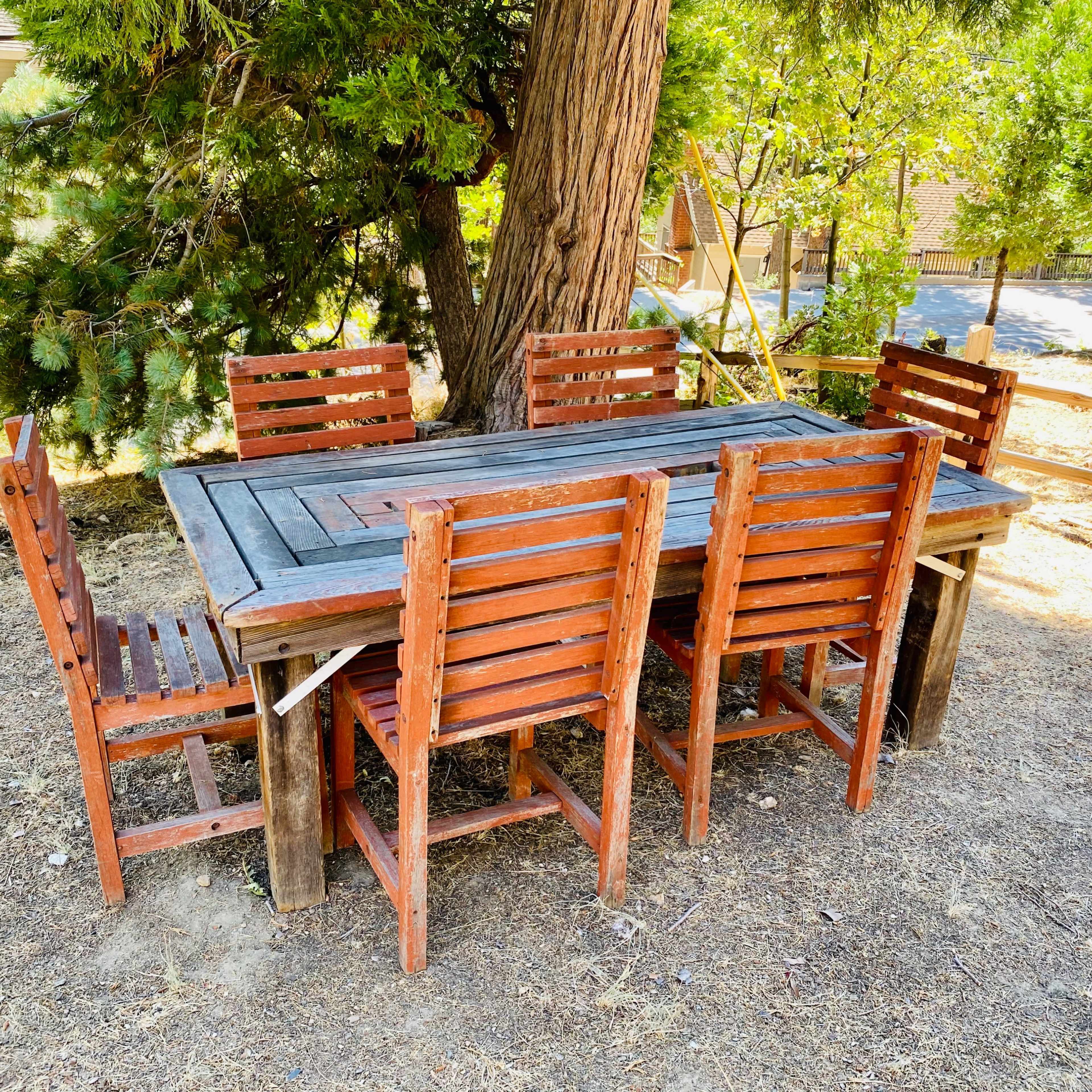 A wooden outdoor dining table surrounded by six matching chairs sits on bare ground under a tree.