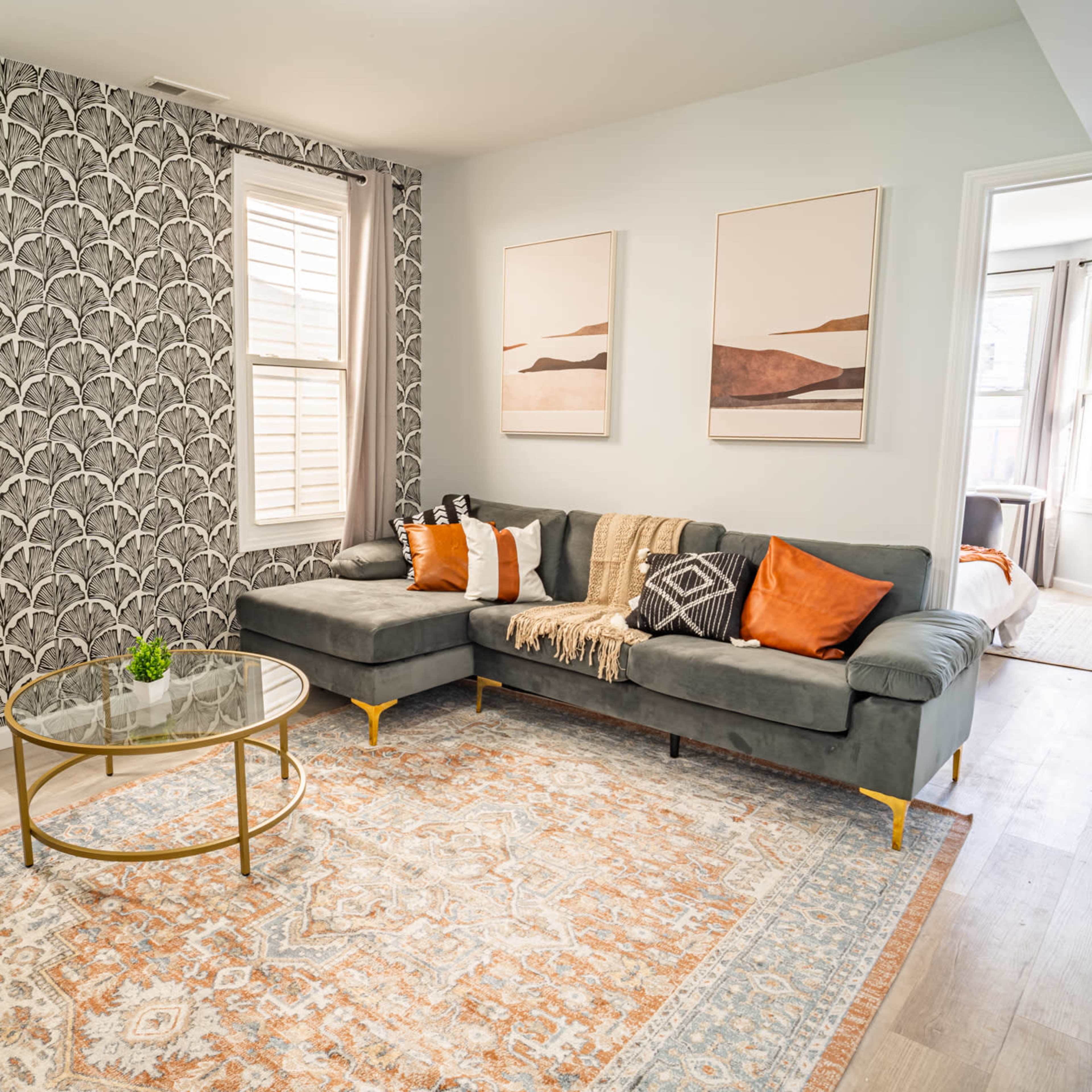 A modern living room featuring a gray sectional sofa, a round glass coffee table, decorative artwork on the walls, and large windows allowing natural light.