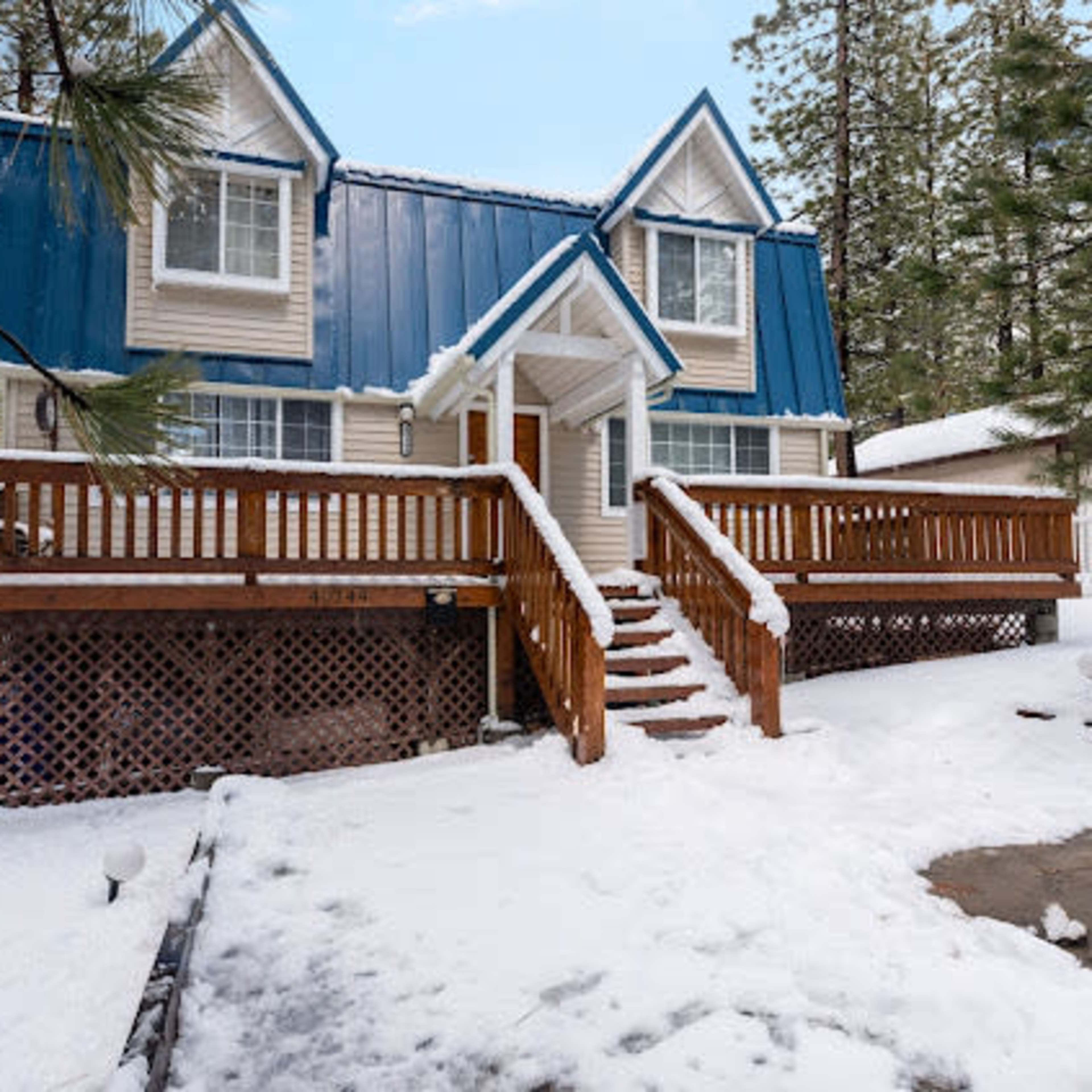 A two-story house with a blue metal roof and wooden deck is surrounded by snow and pine trees.