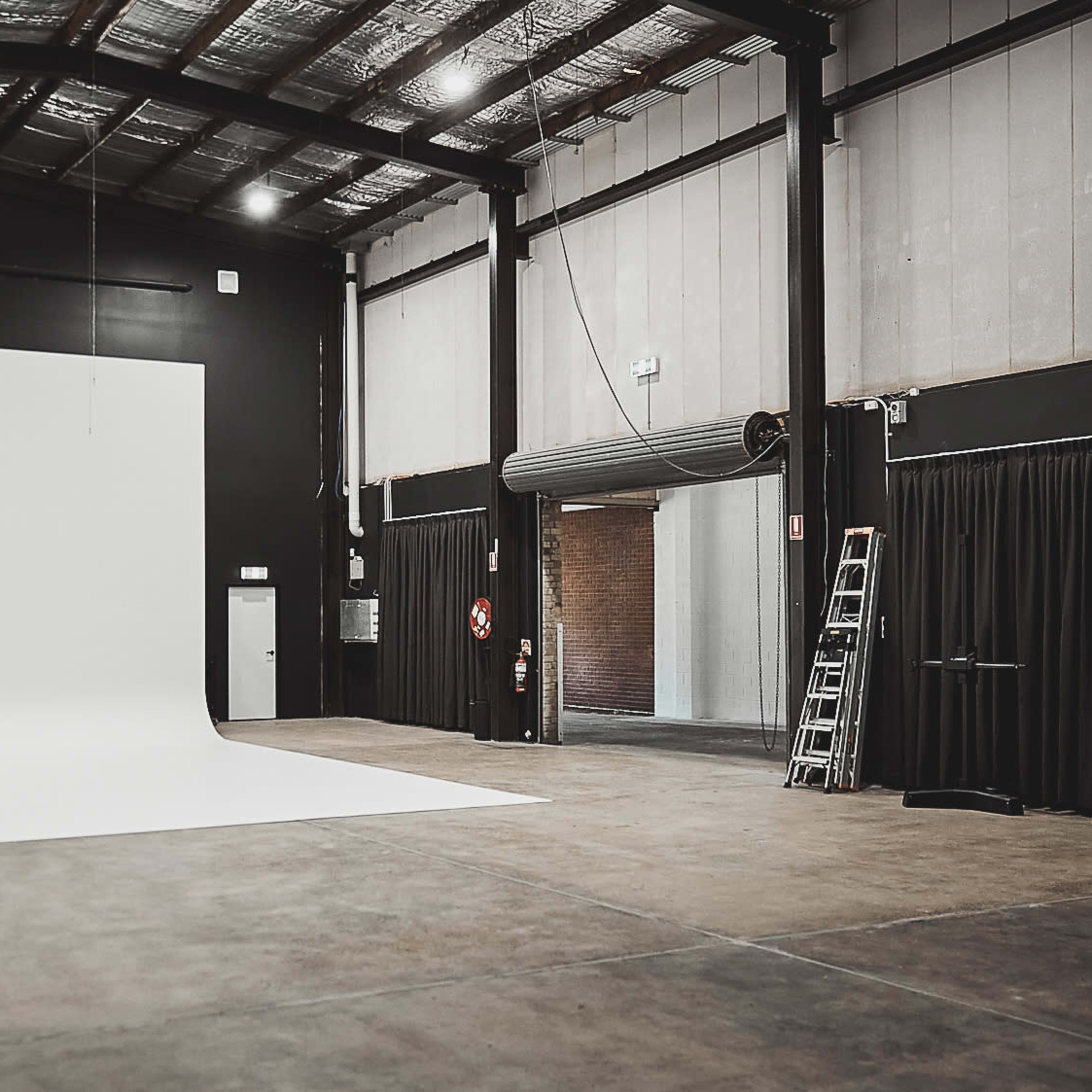 A spacious photography studio featuring a large white backdrop, black walls, and industrial lighting fixtures.