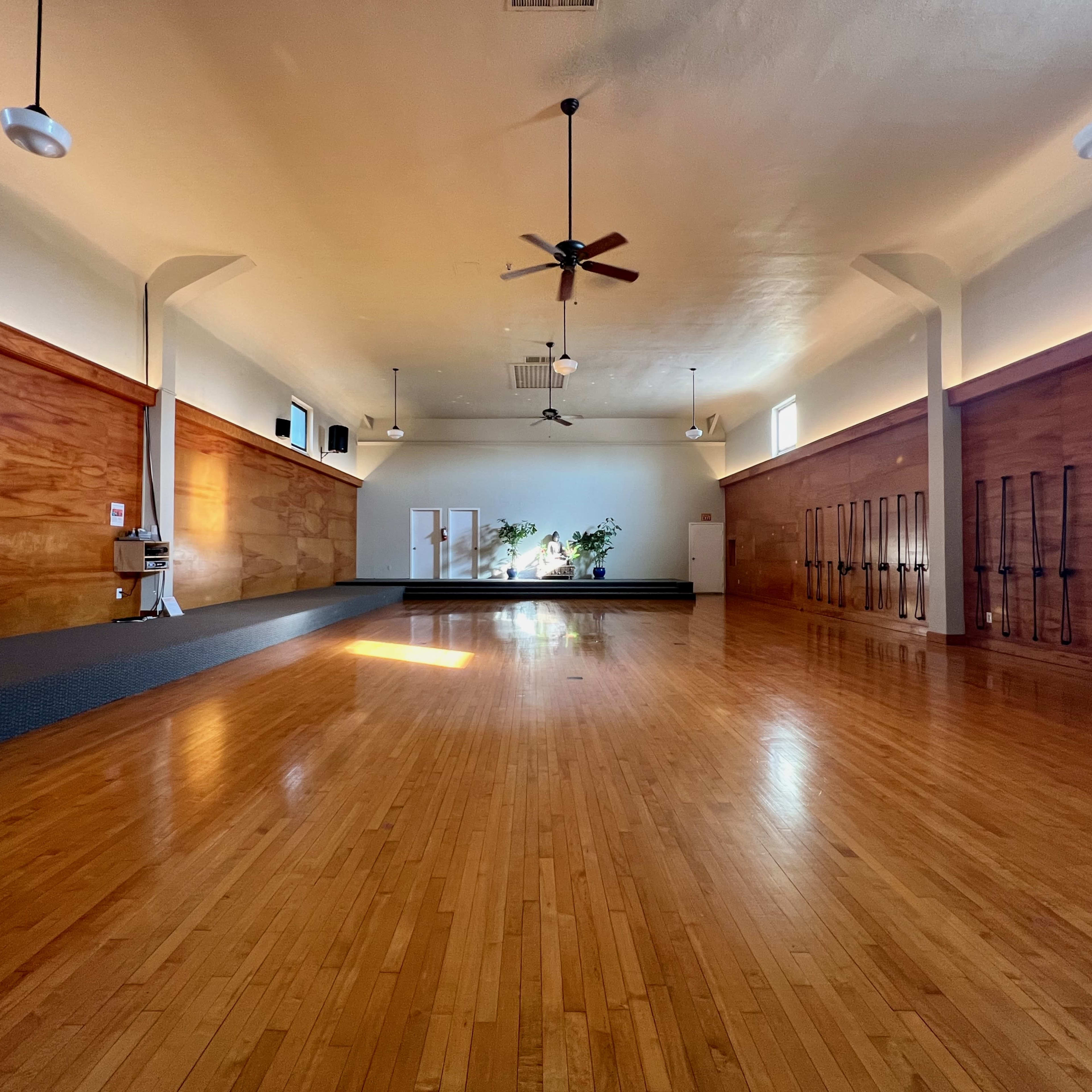 An empty yoga studio with wooden floors, large windows letting in natural light, and wall-mounted yoga props.