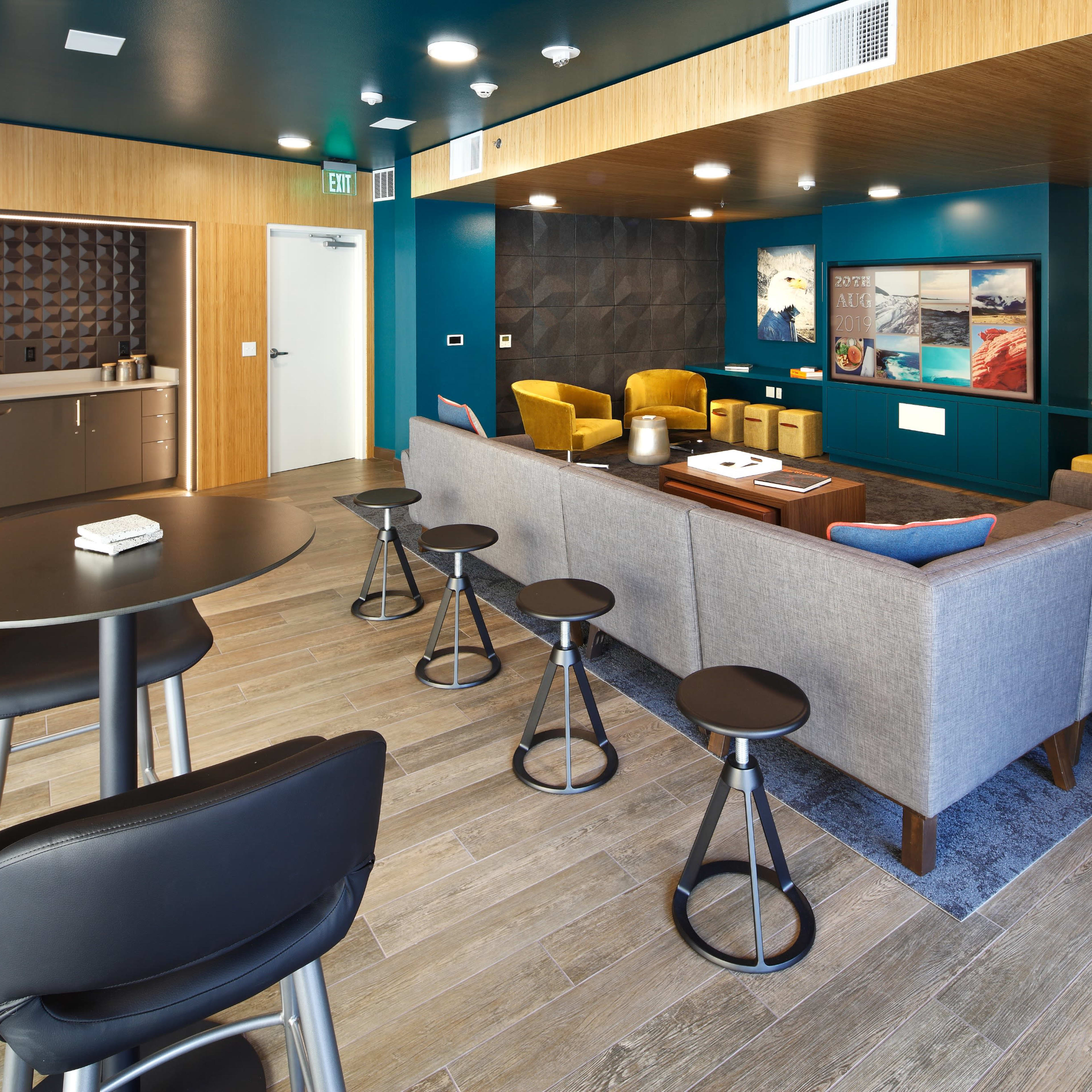 A modern lounge area featuring a gray sectional sofa, yellow armchairs, a coffee table, and a bar counter with high stools against a wooden wall.