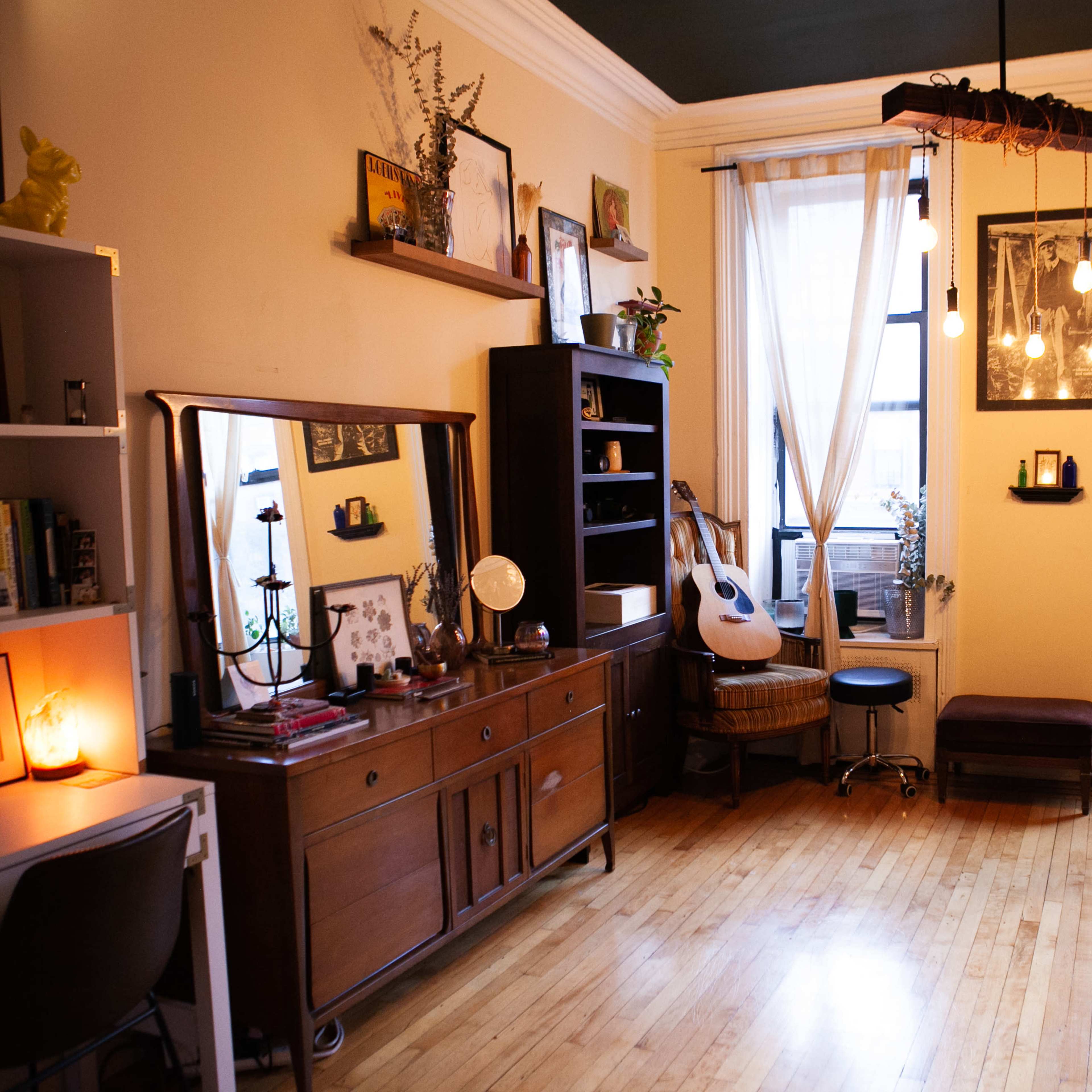 An interior room with wooden floors, a large window, plants, a guitar, and various pieces of furniture arranged throughout.