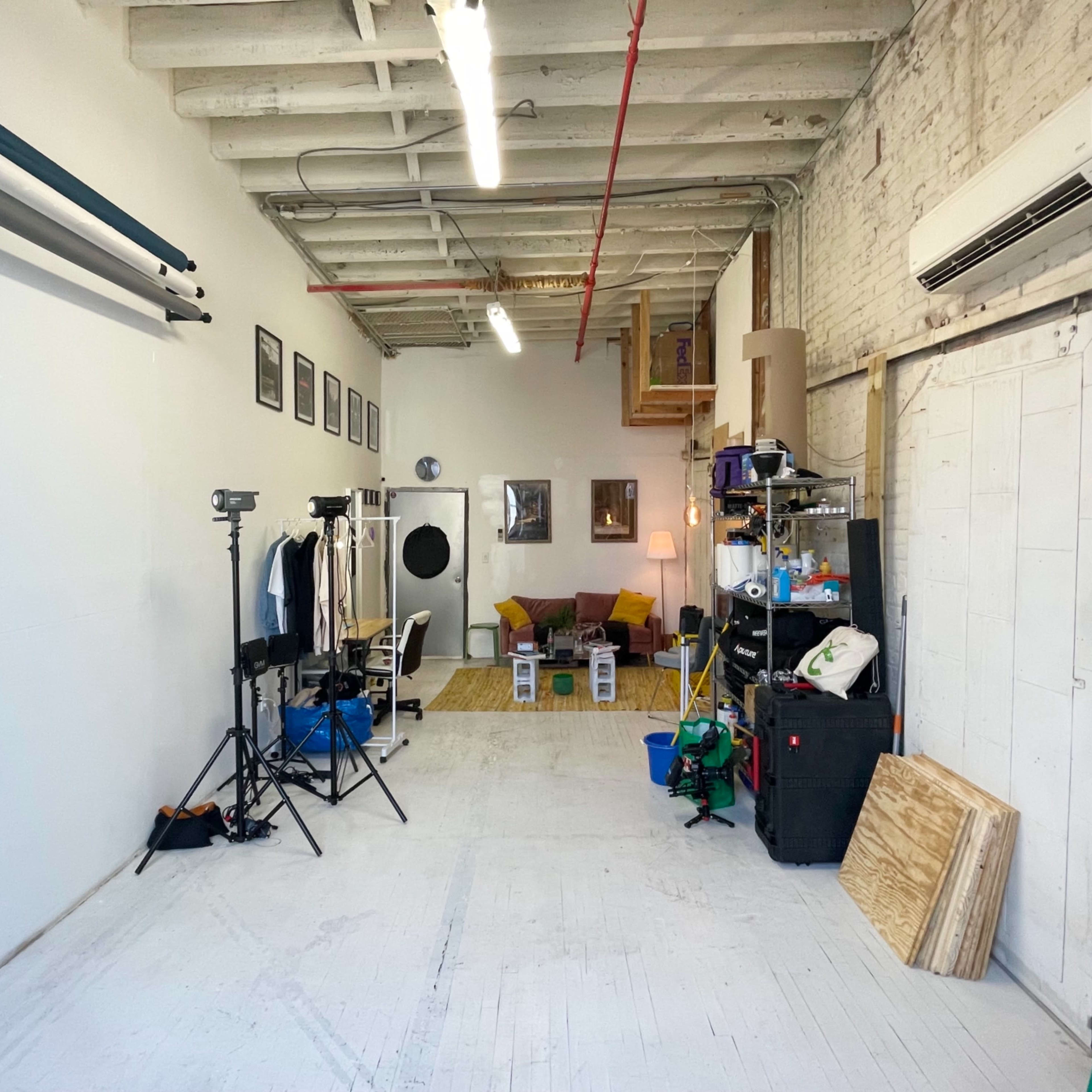 An empty studio space with equipment, furniture, and storage items along one side, and white walls and a wood floor.