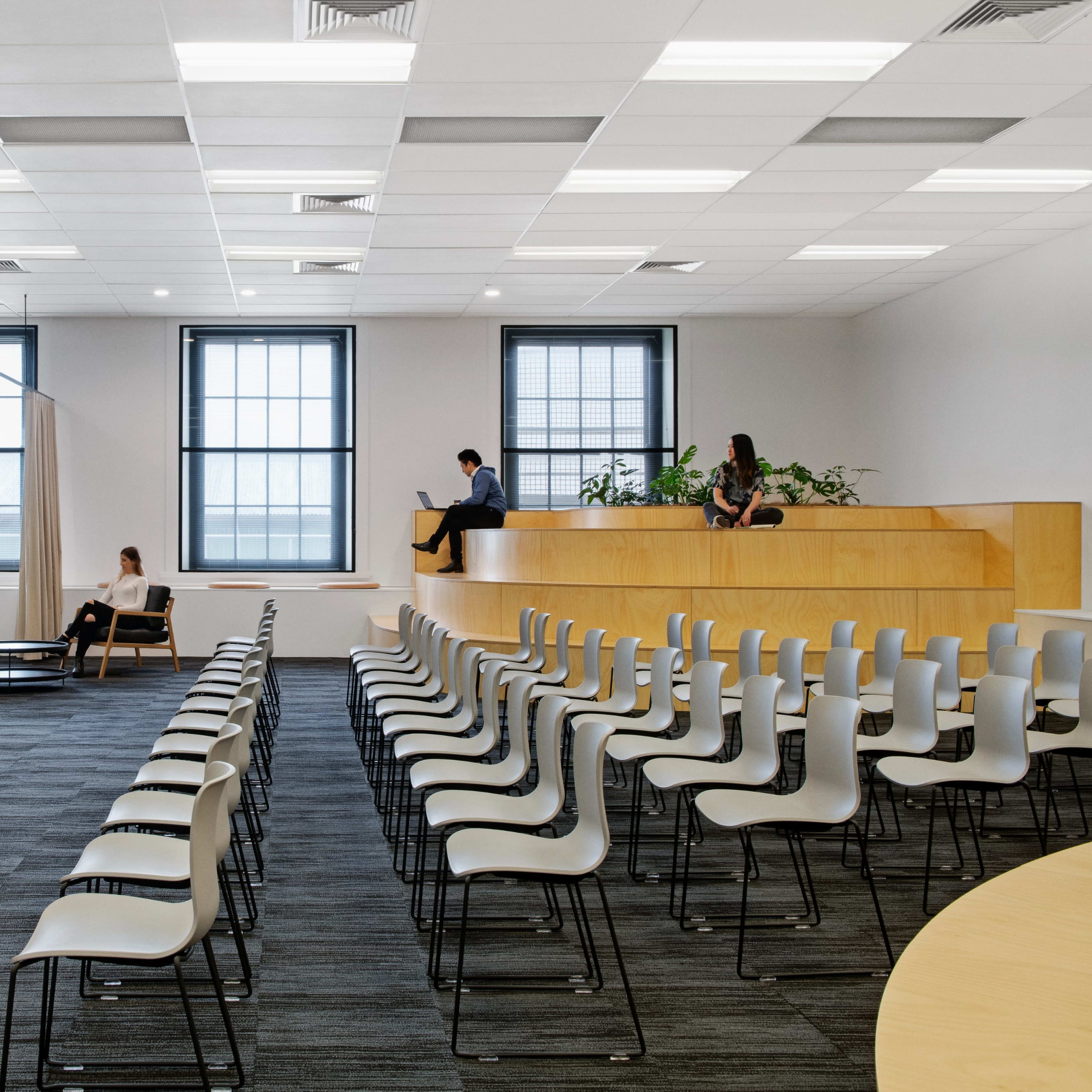 A modern, minimalist office space with rows of chairs facing a large wooden panel that houses a TV, complemented by seating areas and large windows.