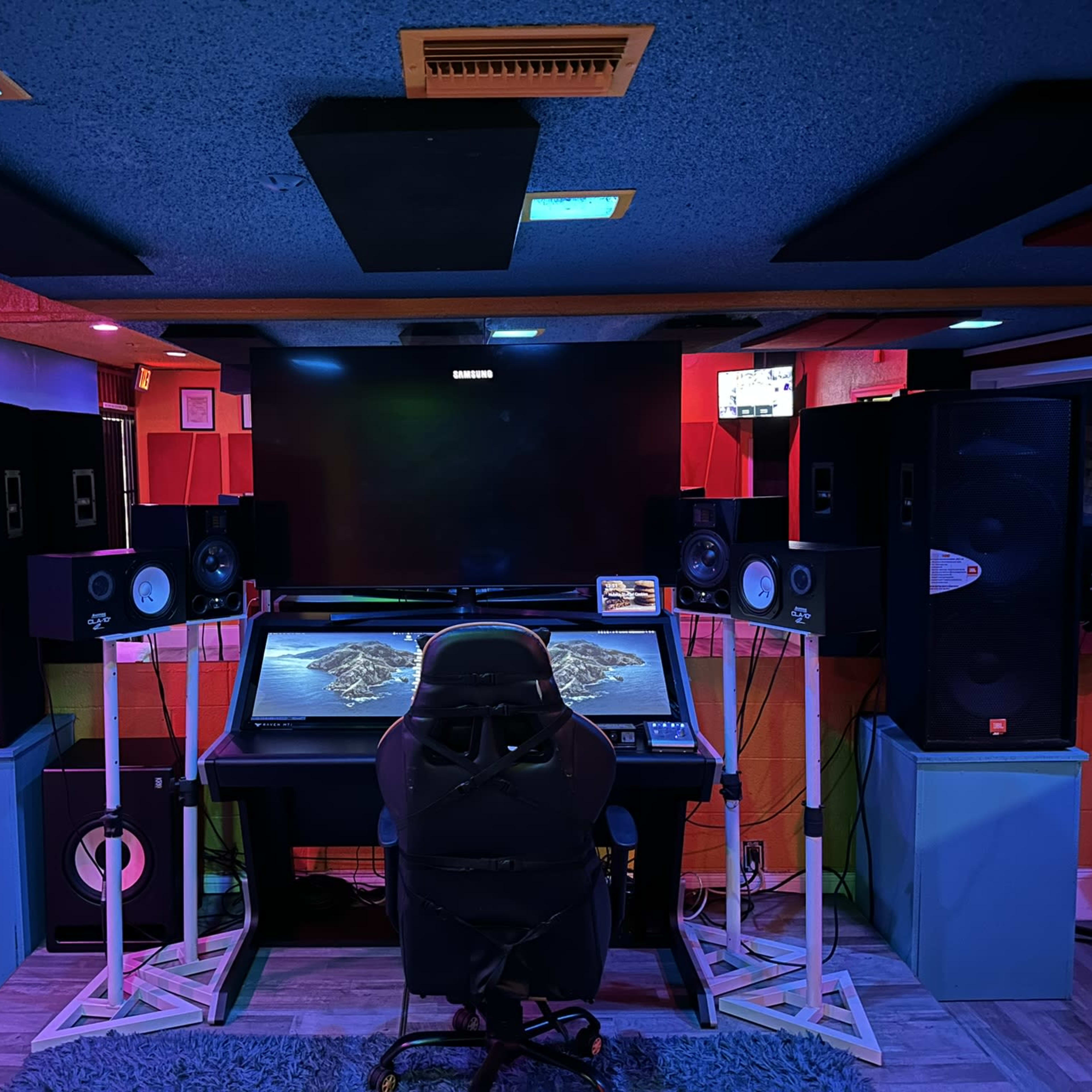 A music studio with a central mixing desk, large speakers on either side, and a television mounted above.