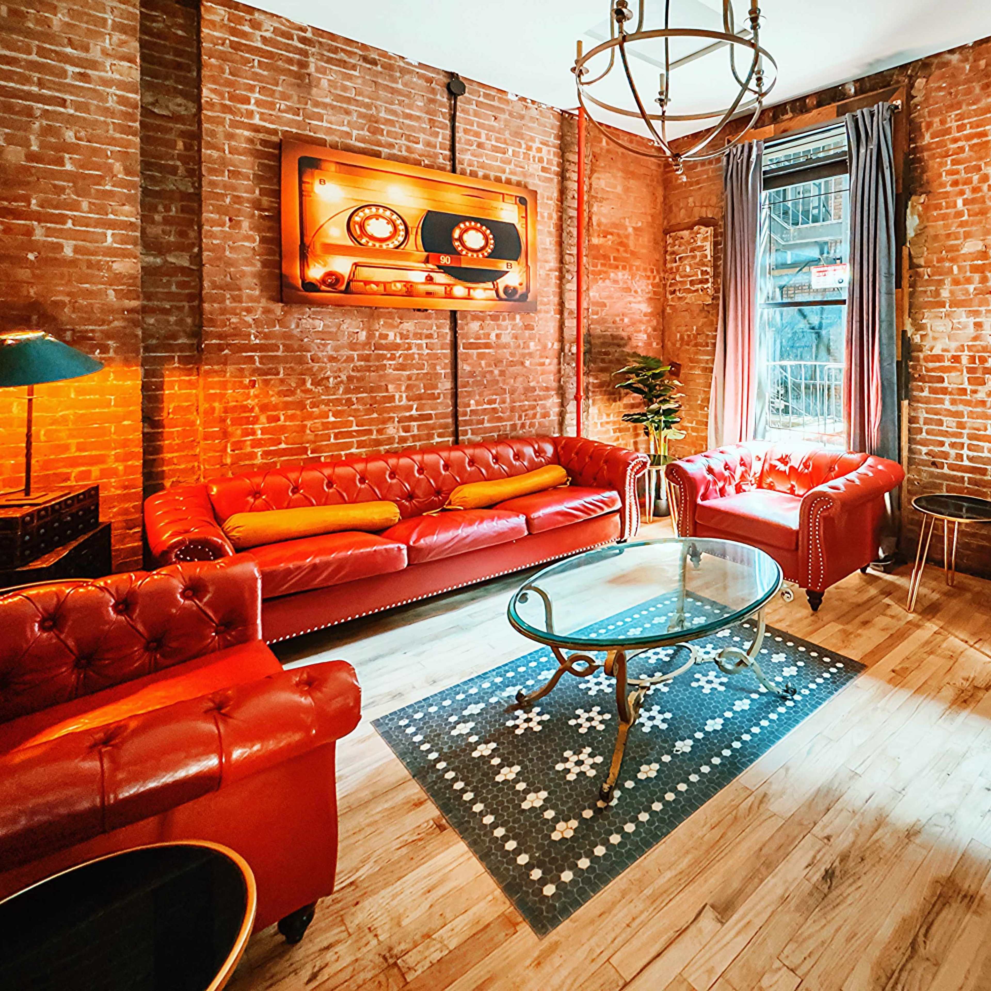 A cozy room with exposed brick walls, featuring red leather sofas, a blue armchair, a glass coffee table, and vibrant artwork on the walls.