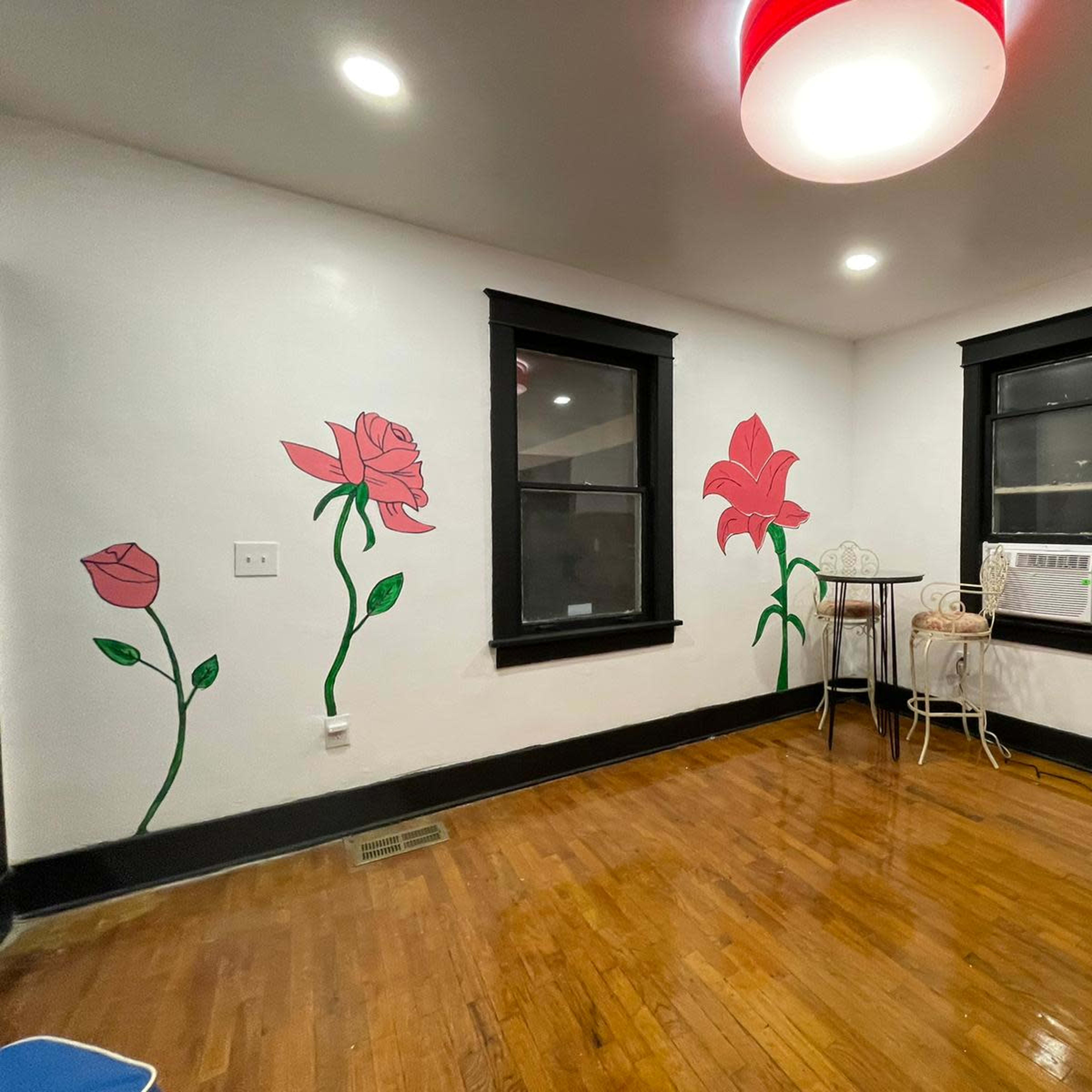 The room features hardwood floors, black window frames, and painted flower murals on the white walls, along with a small table and an air conditioning unit.